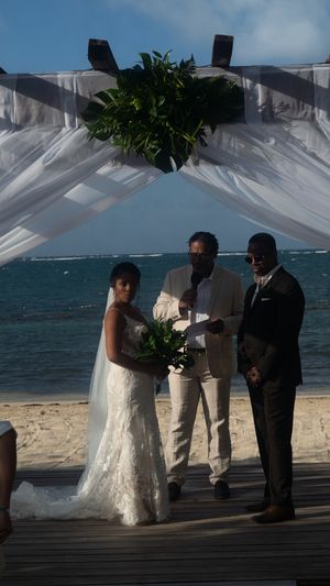 A beachside wedding ceremony takes place under a white draped canopy with turquoise ocean waters and blue sky in the background.