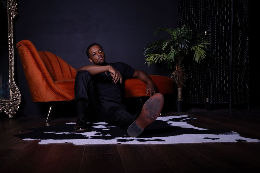 Man in black outfit relaxing on red velvet sofa with cowhide rug and tropical plant in dark studio.