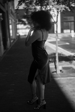 A figure in a black dress and heels stands in dramatic shadow on a sidewalk, with a natural afro hairstyle silhouetted.