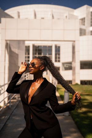 Joyful woman in black blazer holding braided hair and sunglasses while standing outdoors on sunny day.