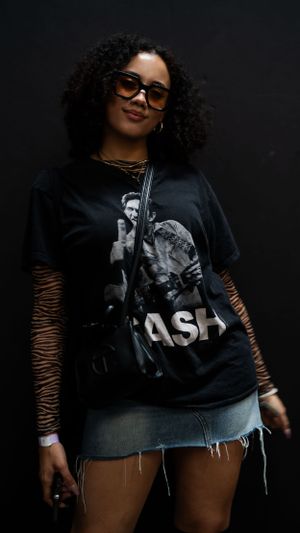 Woman with curly hair wearing sunglasses and a band t-shirt poses against dark background.