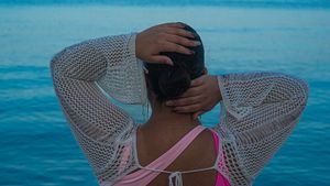 Person wearing a white mesh cover-up and pink swimsuit stands with hands on head against bright blue ocean backdrop.
