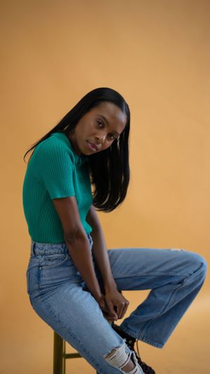 A person in a green ribbed top and distressed jeans poses casually against an orange background.