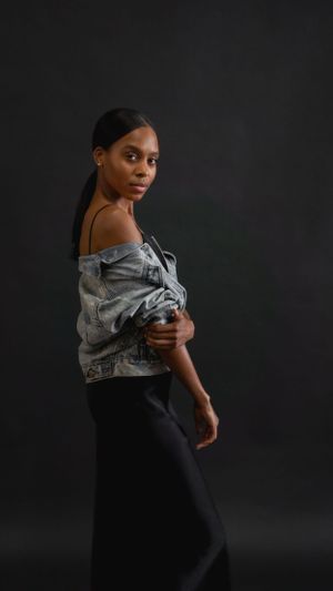 A dramatic fashion portrait against a dark backdrop showing a denim jacket worn off-shoulder with black pants.