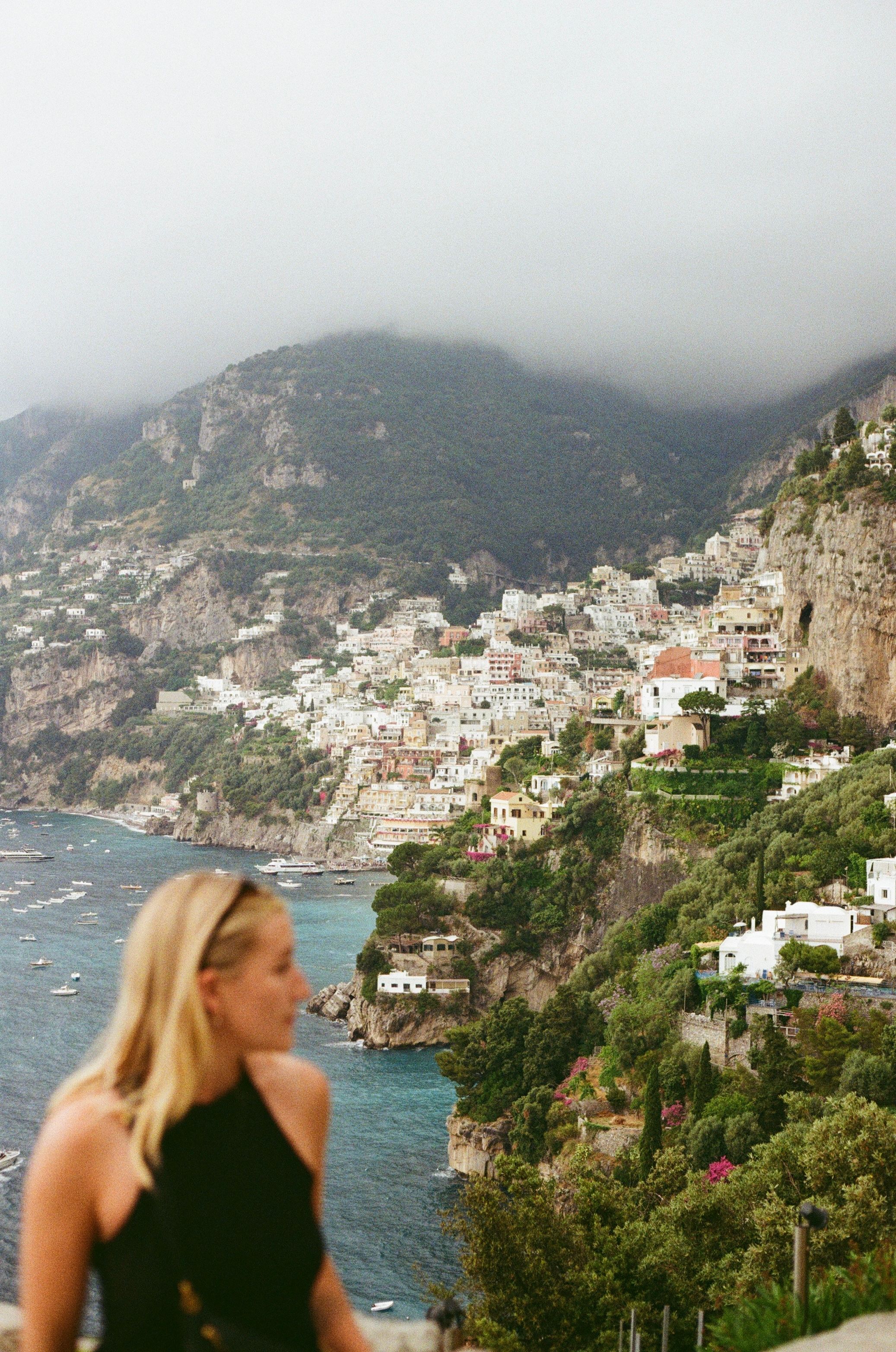 Blonde woman overlooking colorful cliffside village of Positano on the Amalfi Coast, Italy, with misty mountains.