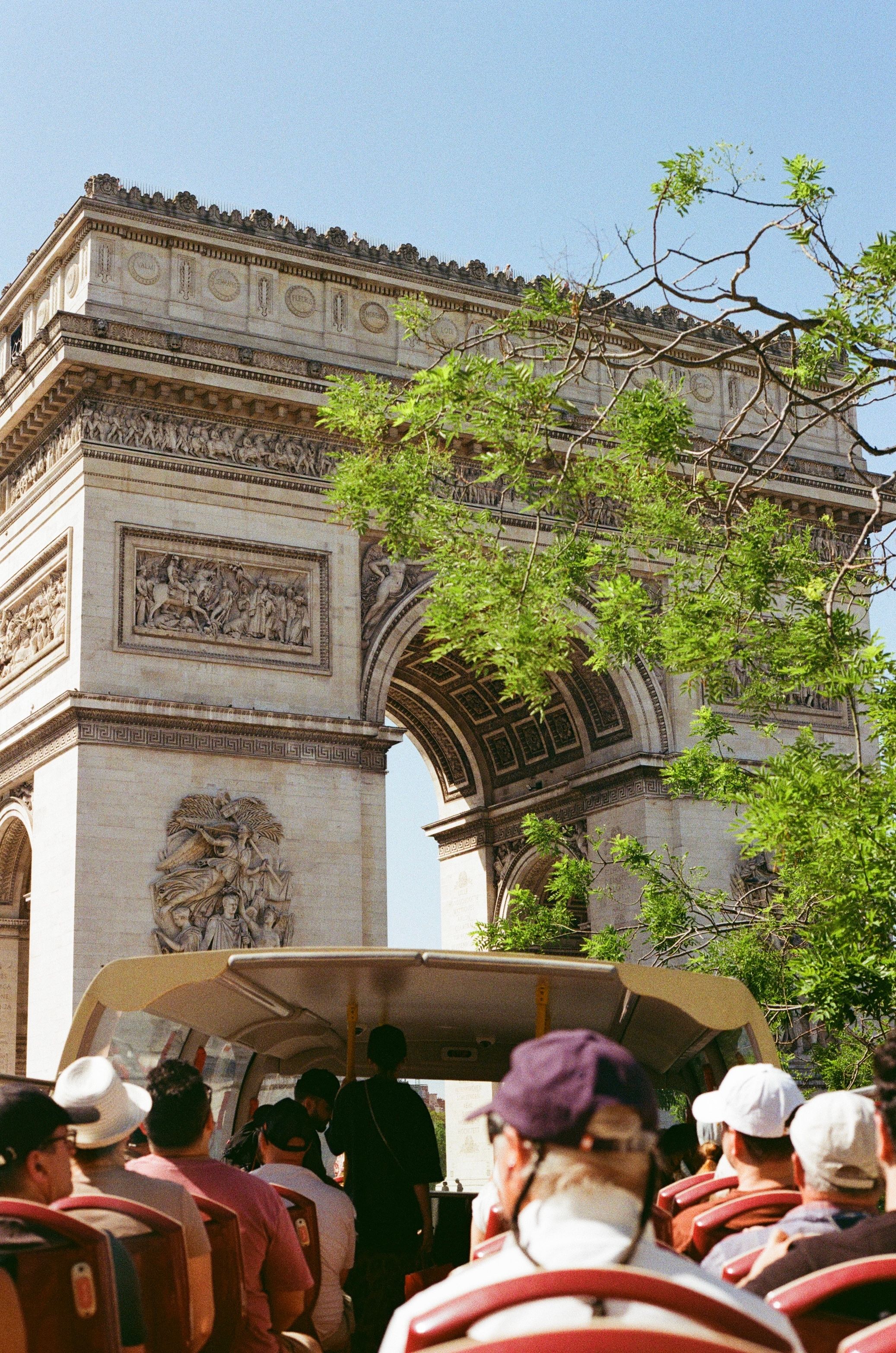 Tourists on open-top bus viewing the Arc de Triomphe in Paris, France, surrounded by green trees under a clear blue sky.