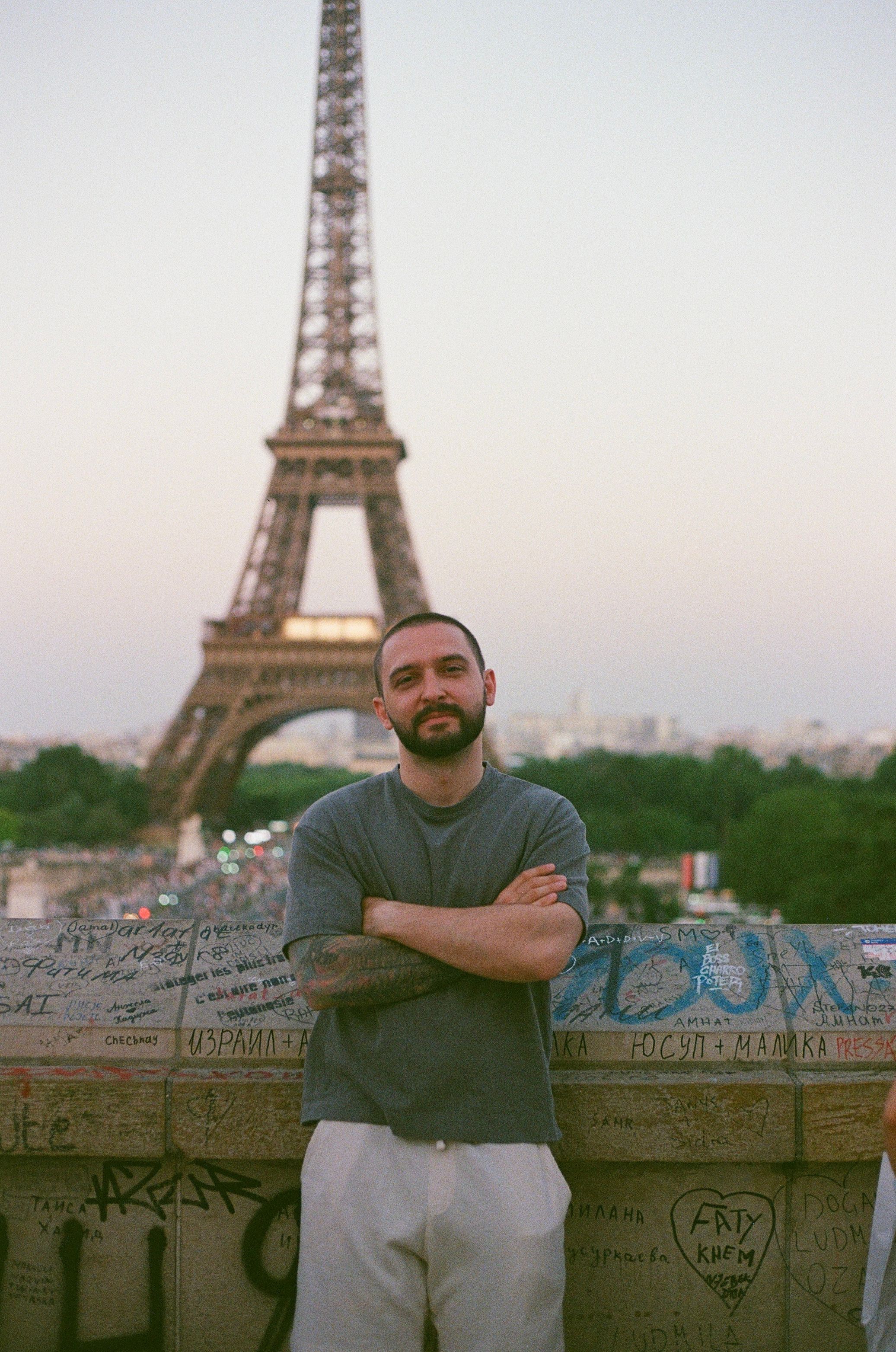 Man with crossed arms standing in front of the Eiffel Tower in Paris at dusk with graffiti wall.