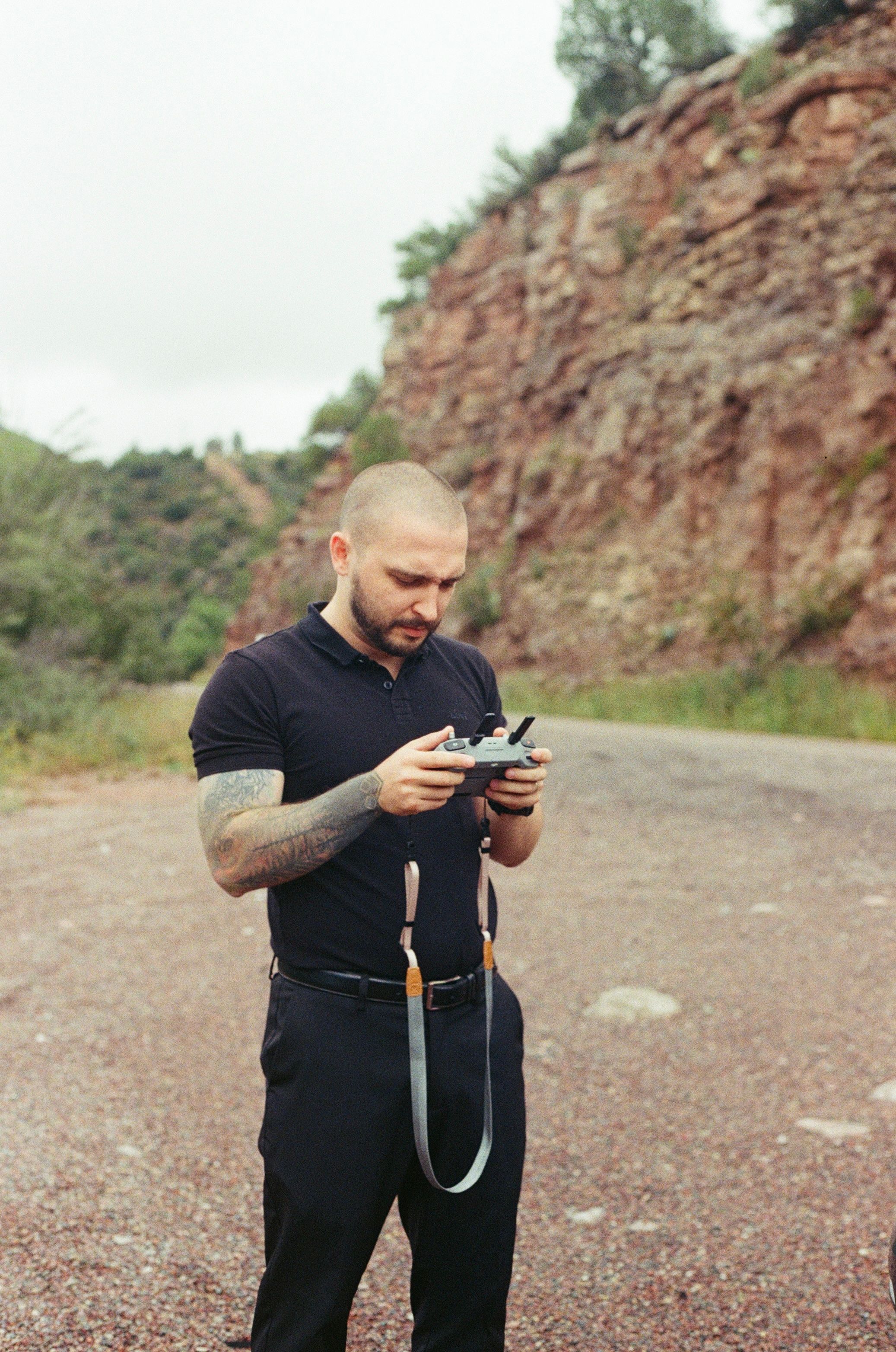Tattooed bald man in black outfit operating a drone remote controller on a rural road with rocky hillside backdrop.