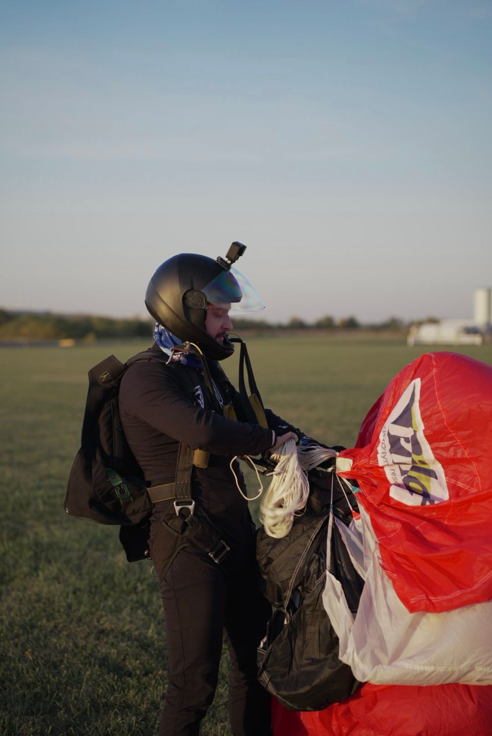 Skydiver in dark gear preparing parachute equipment in an open field at sunset.