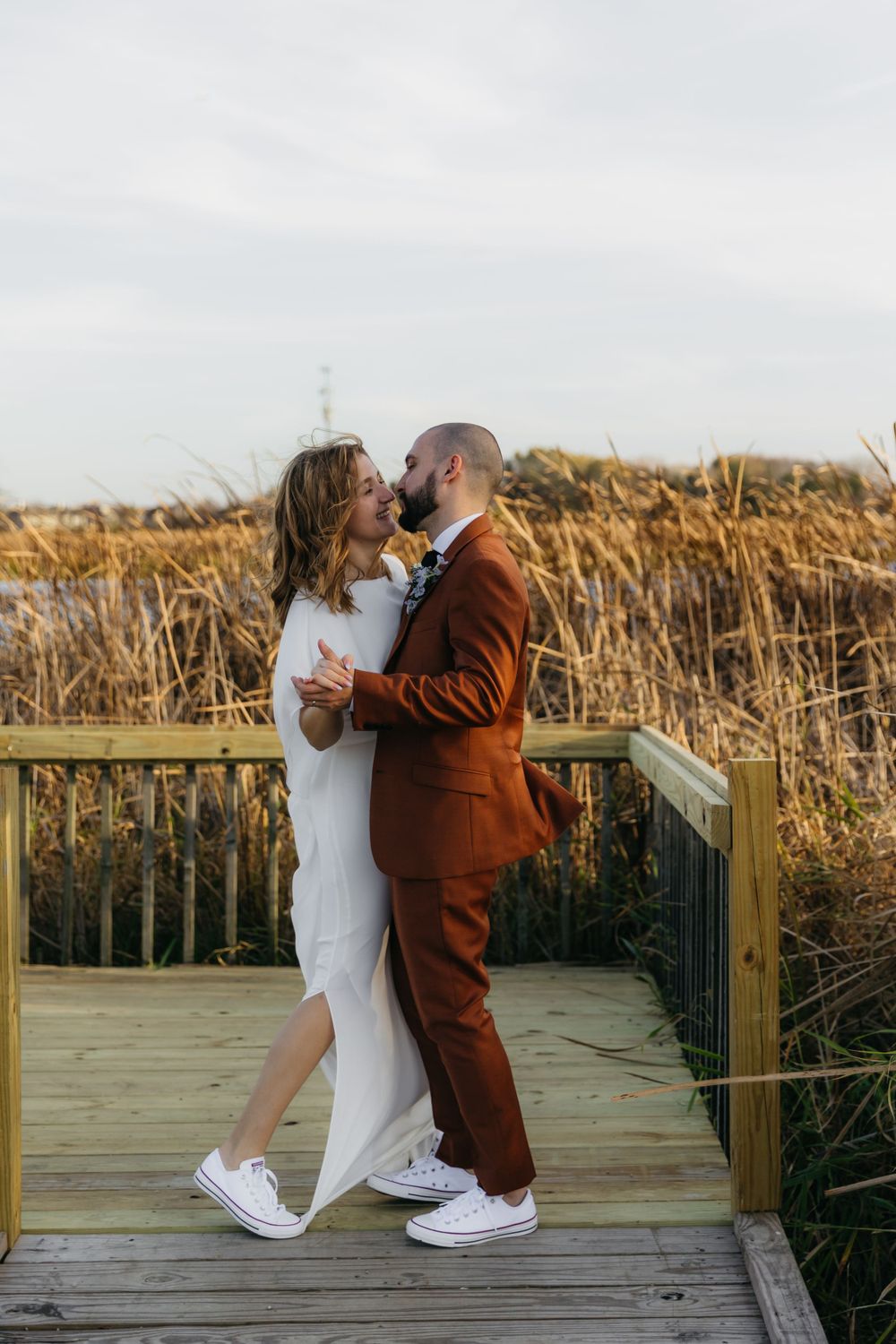 Couple in matching white sneakers sharing a romantic moment on a wooden boardwalk by tall reeds.