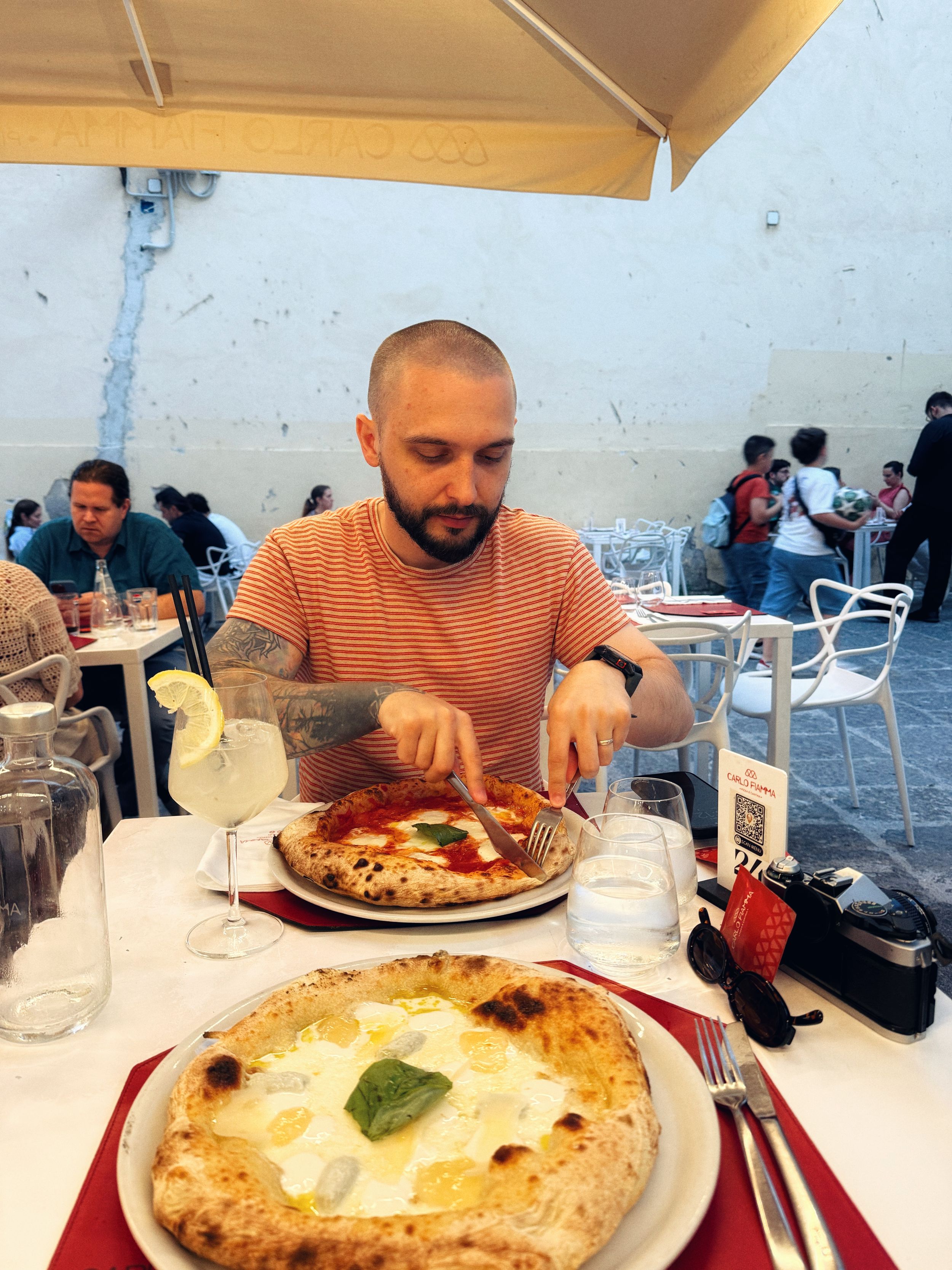Man cutting Neapolitan pizza at outdoor Italian restaurant table with two pizzas, glasses, and diners in background.