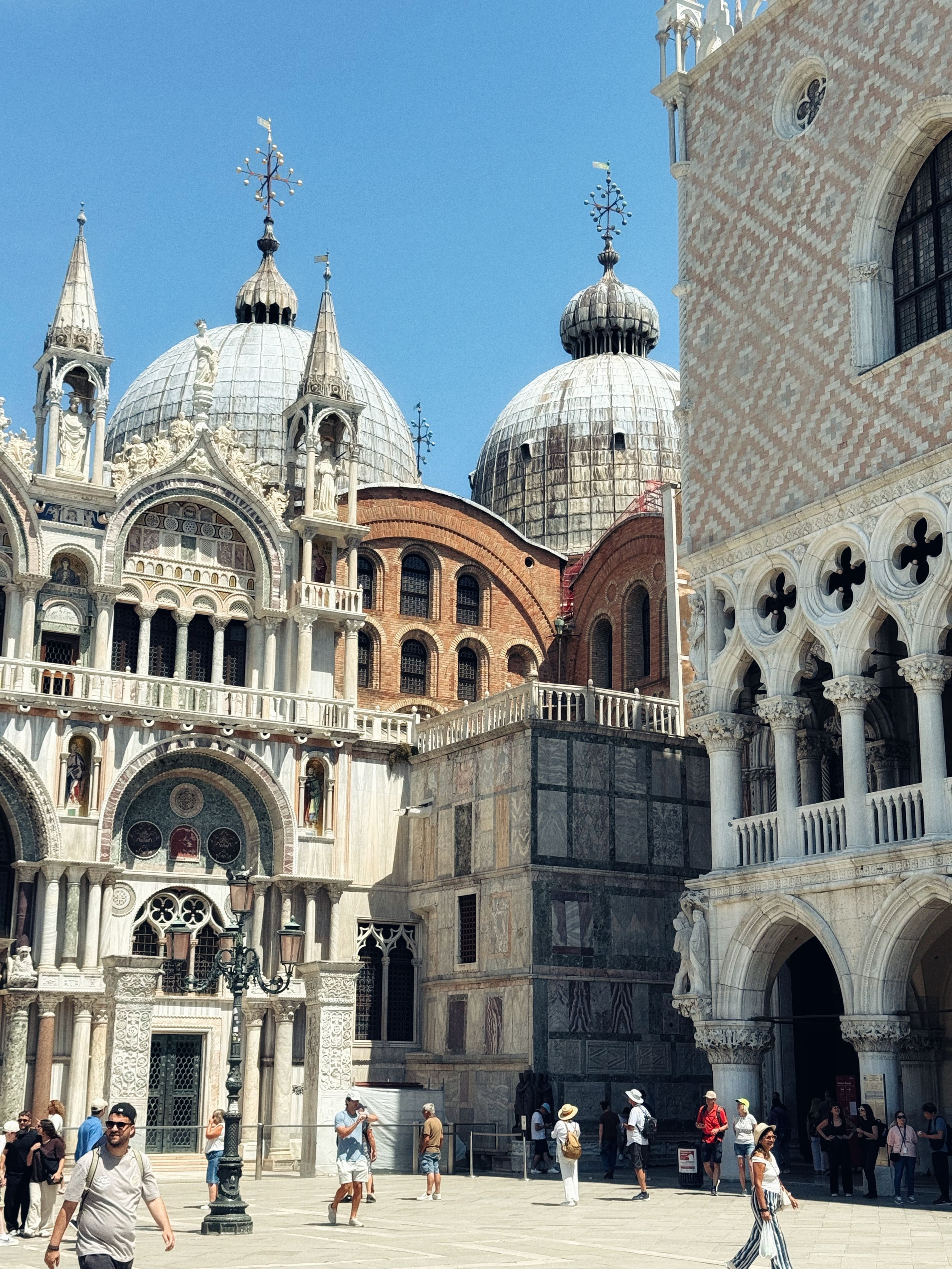 Tourists explore St. Mark's Square in Venice, Italy, with the ornate Byzantine Basilica and Doge's Palace under clear blue skies.