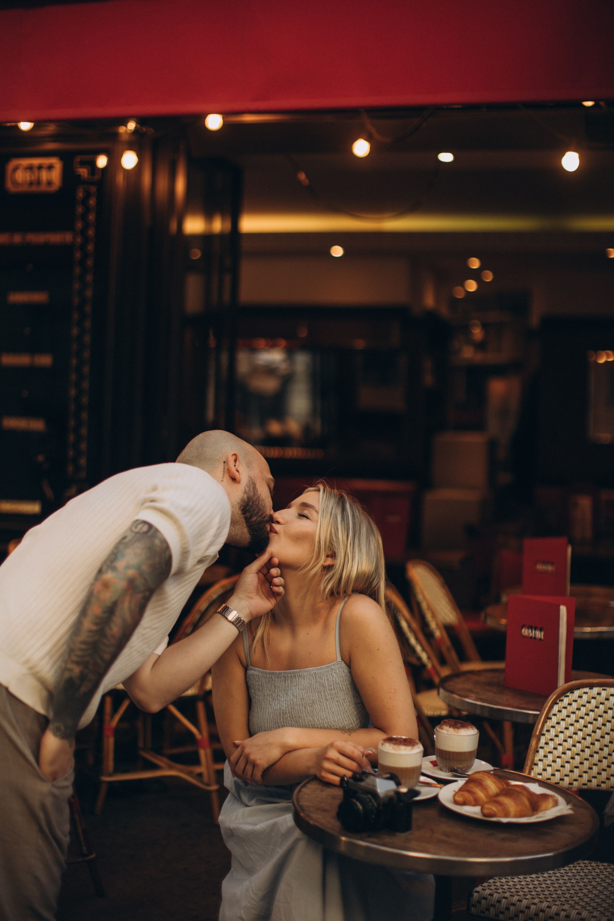 A tattooed man kisses a smiling blonde woman at a Parisian cafe table with croissant and coffee.