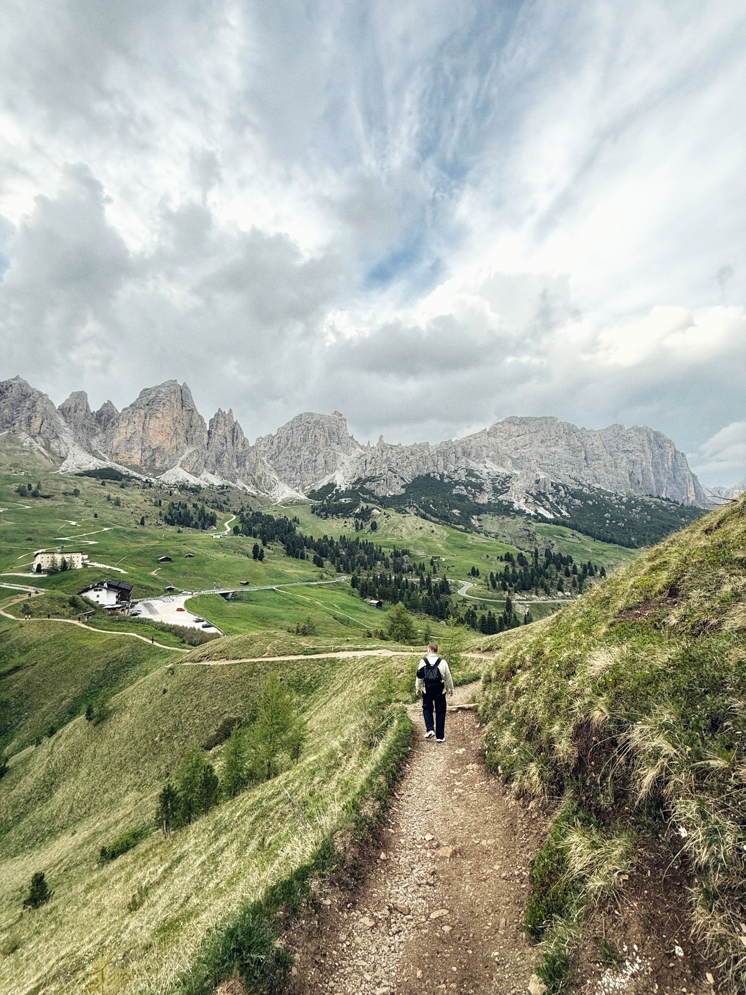 Hiker walking along a narrow mountain trail in the Dolomites with green valleys and rocky peaks under a cloudy sky.