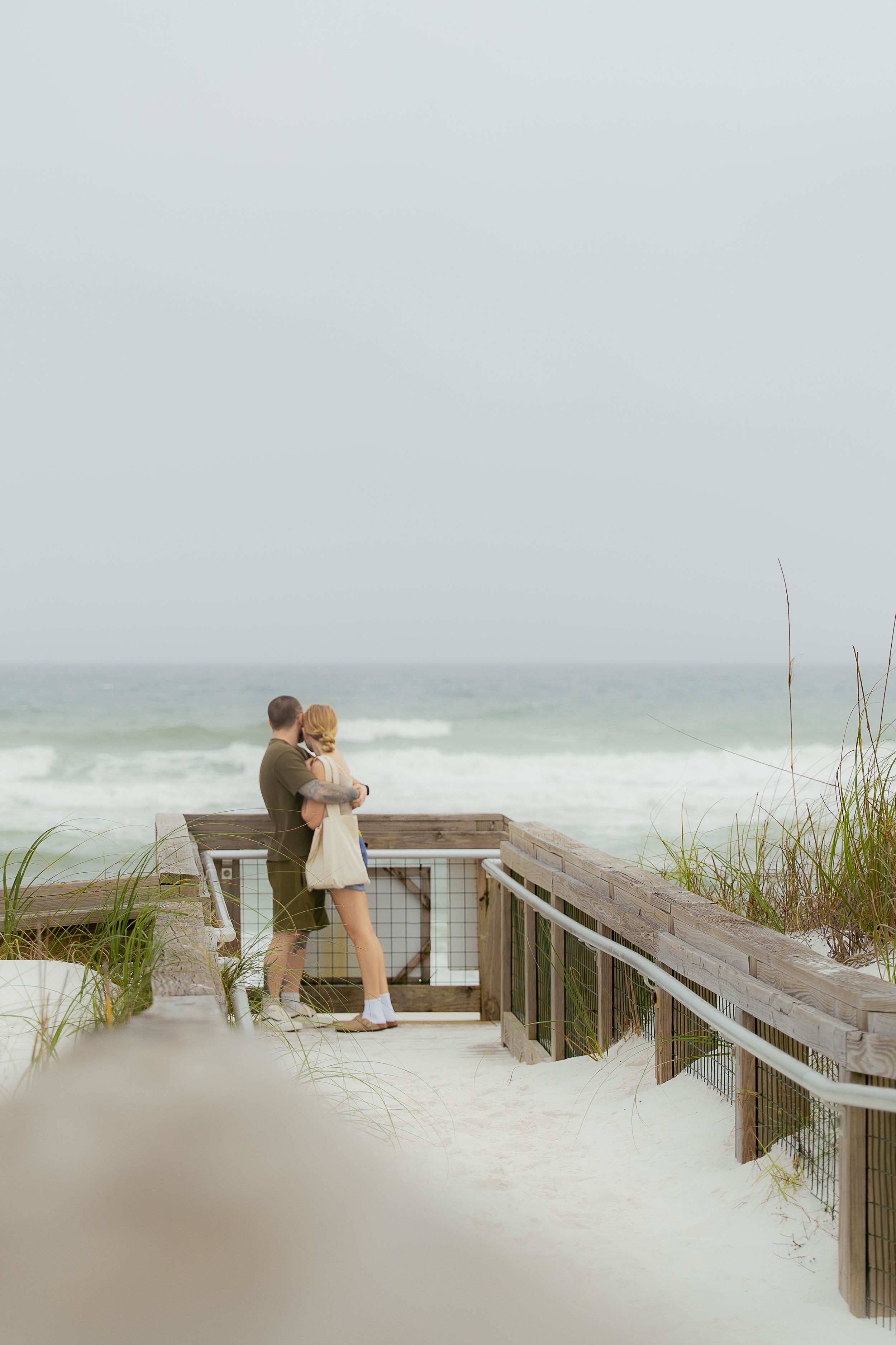 A couple embraces on a wooden boardwalk overlooking a foggy beach with sea grass and waves in the background.