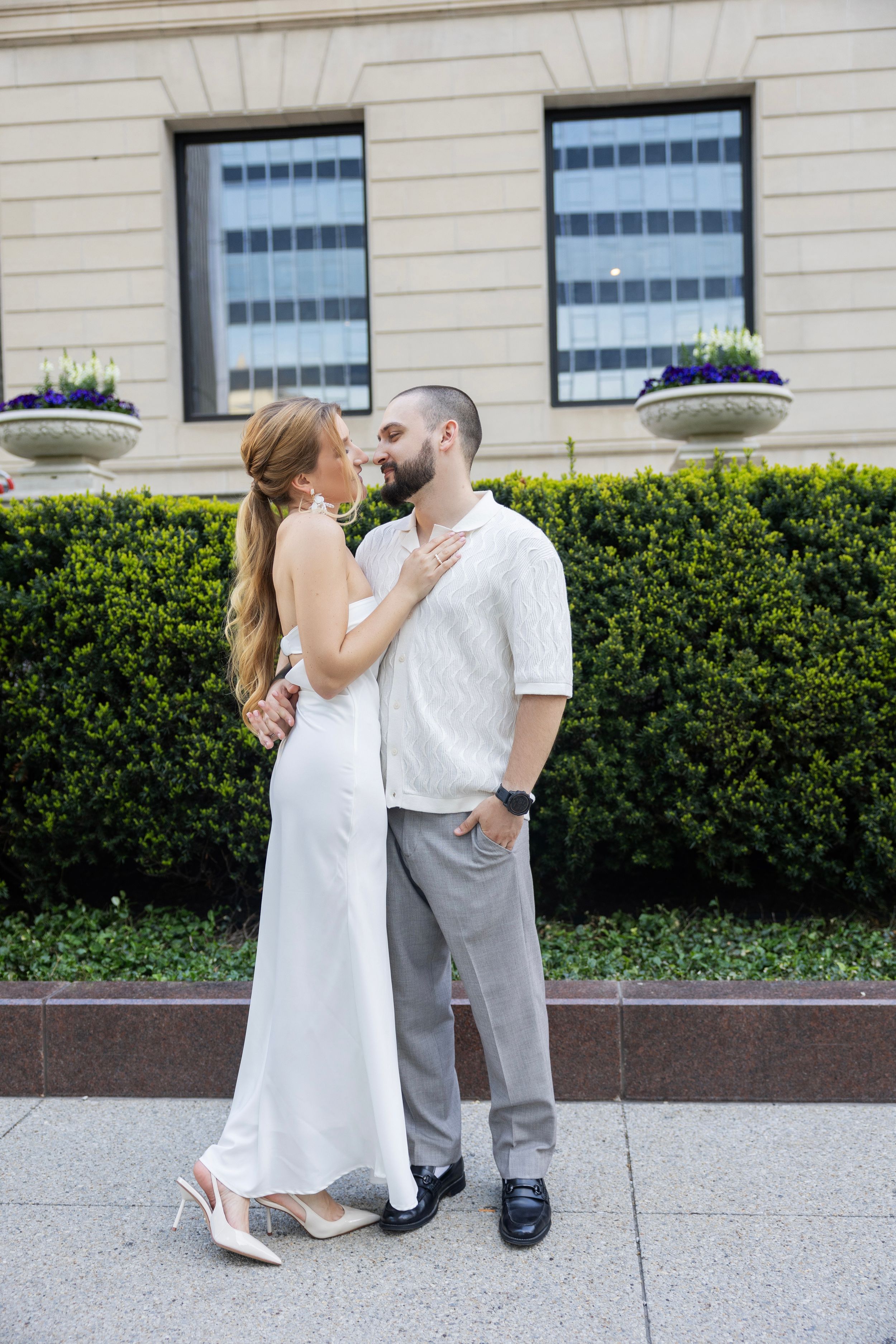A couple shares an intimate moment in front of a building with manicured hedges and hanging flower baskets.
