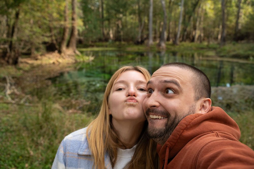 Couple taking selfie in front of scenic forest pond.
