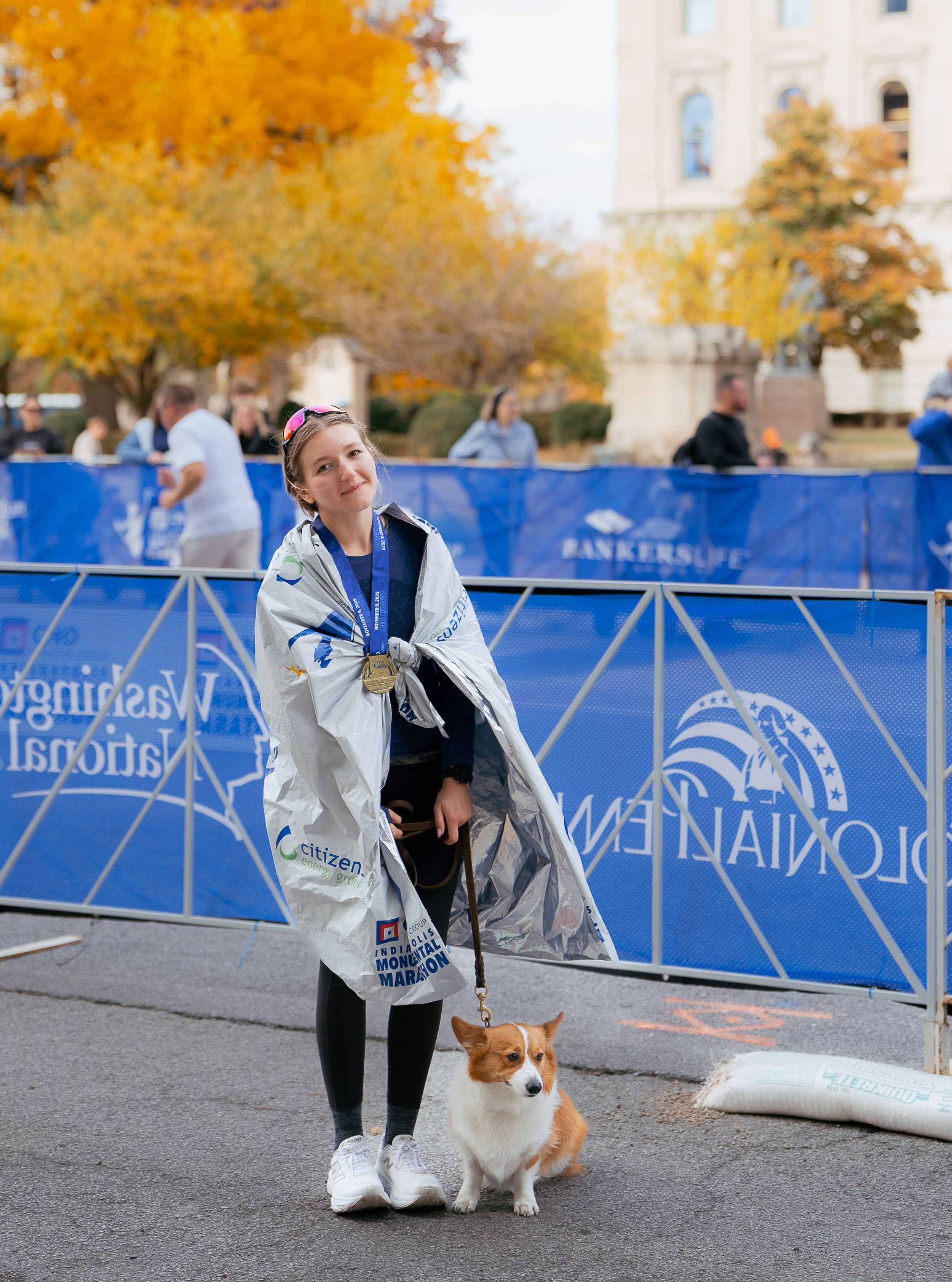 A Corgi dog and owner interact at an outdoor event with blue barriers and autumn trees in the background.