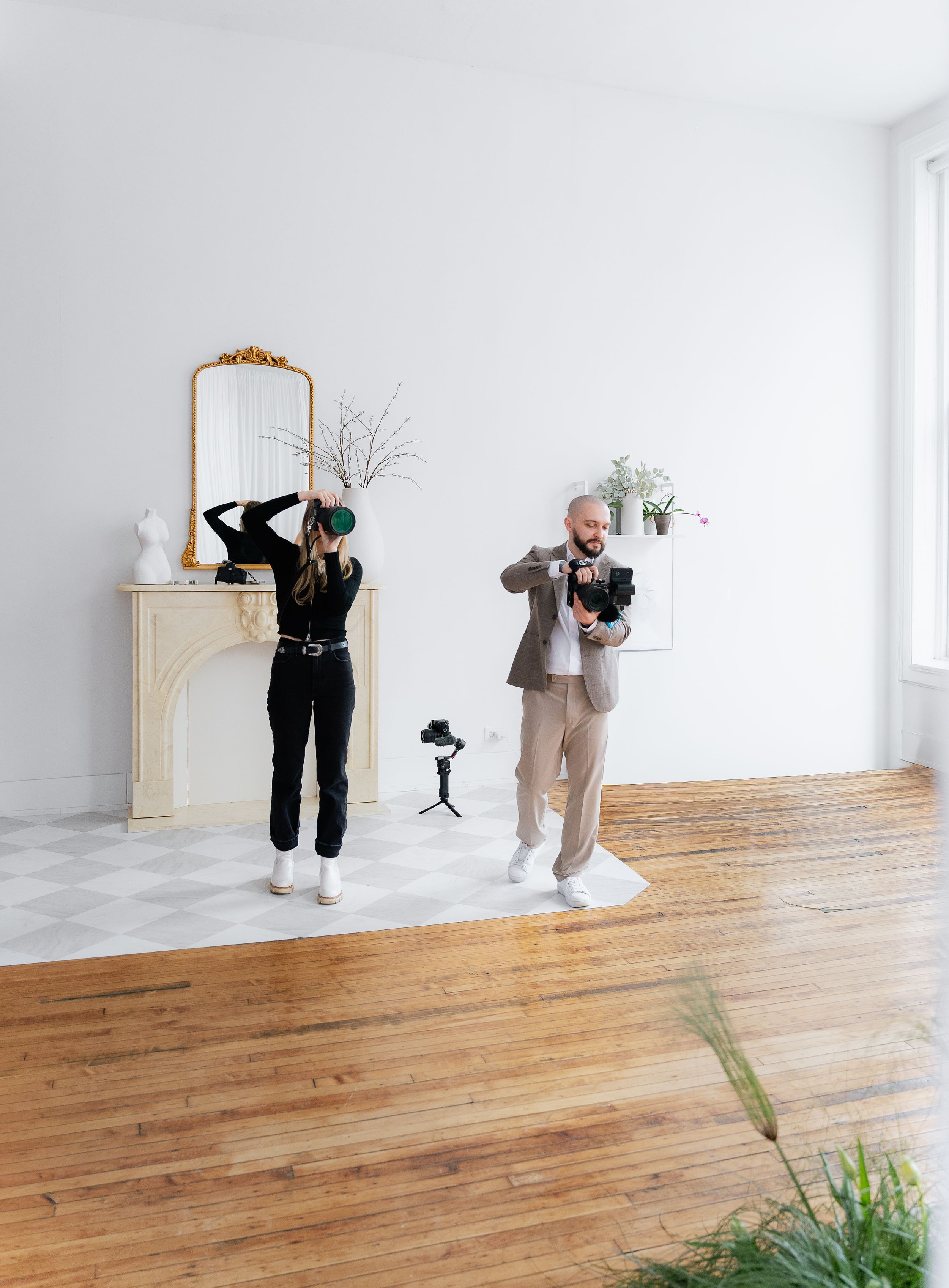 Two photographers in contrasting outfits pose playfully in a minimalist white room with a decorative fireplace mantel.