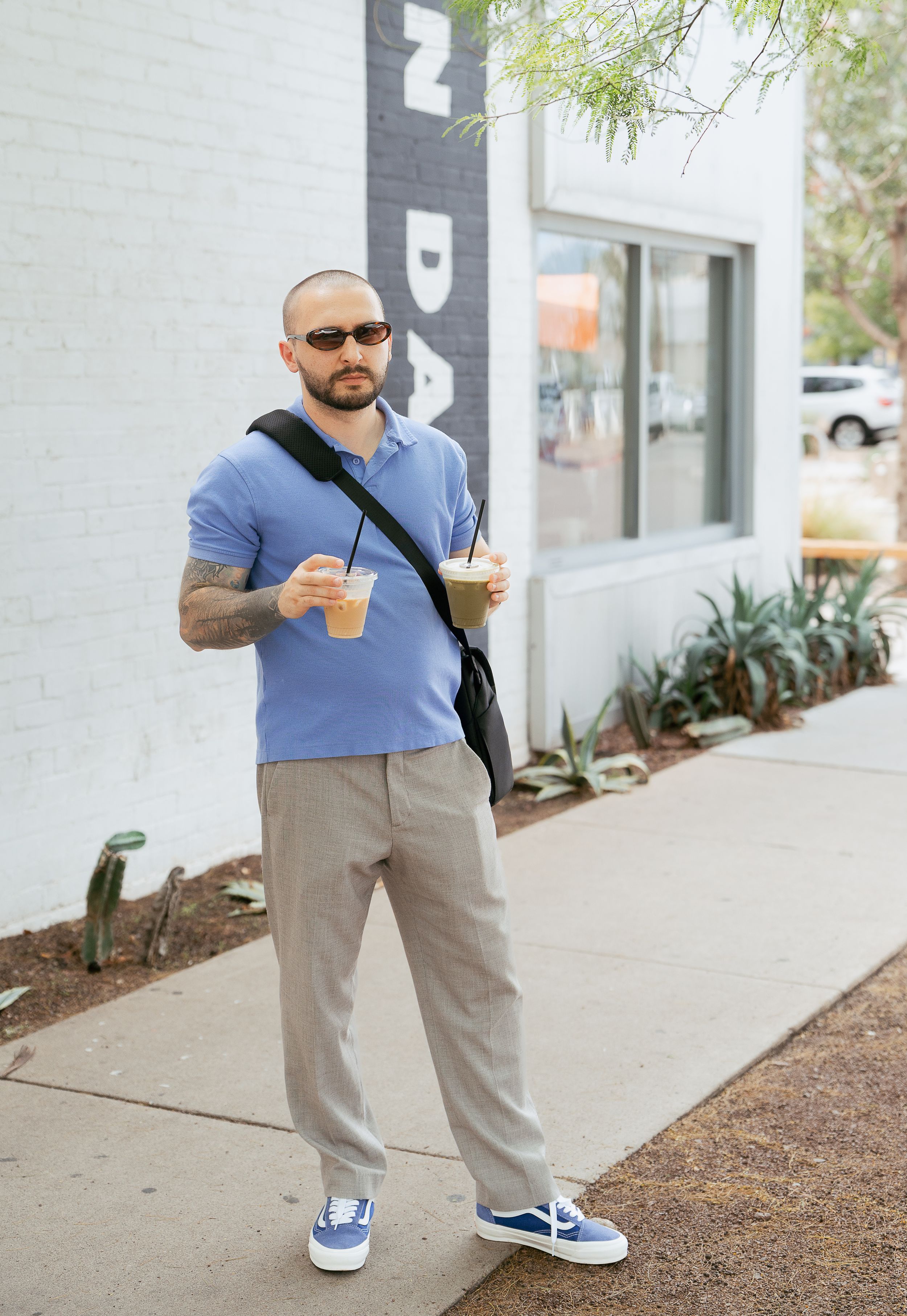 Someone in a blue shirt and khaki pants stands outside a building while holding an orange drink.