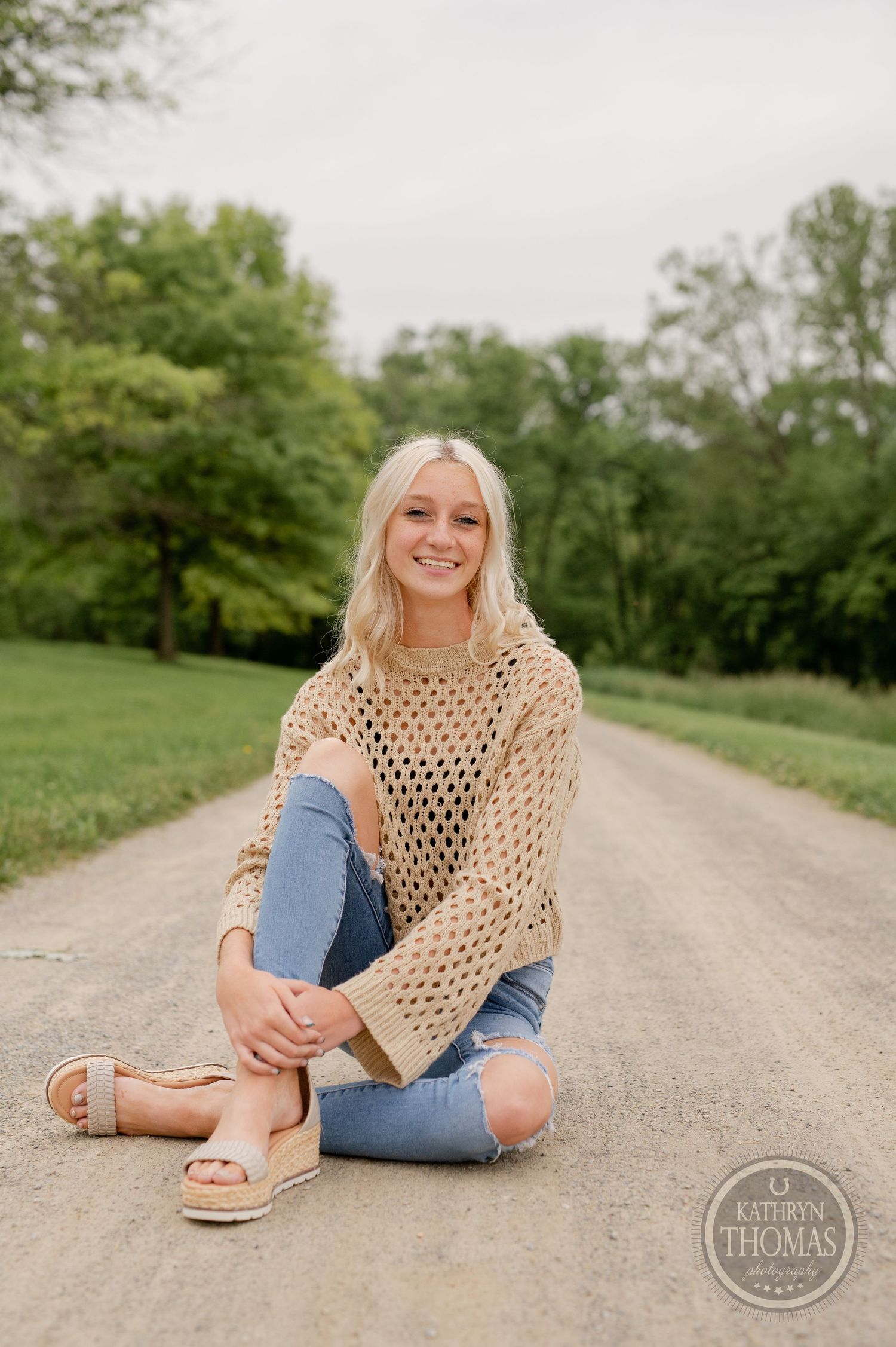 Casual seated portrait on a dirt path wearing a tan mesh sweater and destroyed denim.