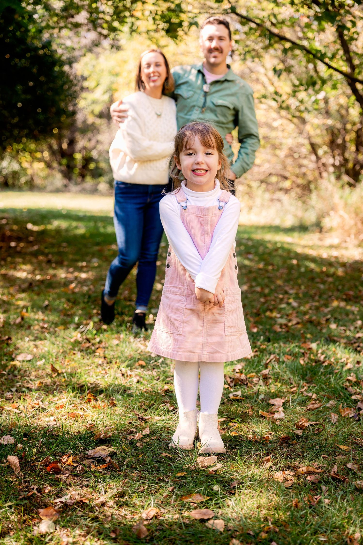 A family stands together on fallen autumn leaves in a park during a sunny outdoor photo session located in Novi Michigan.
