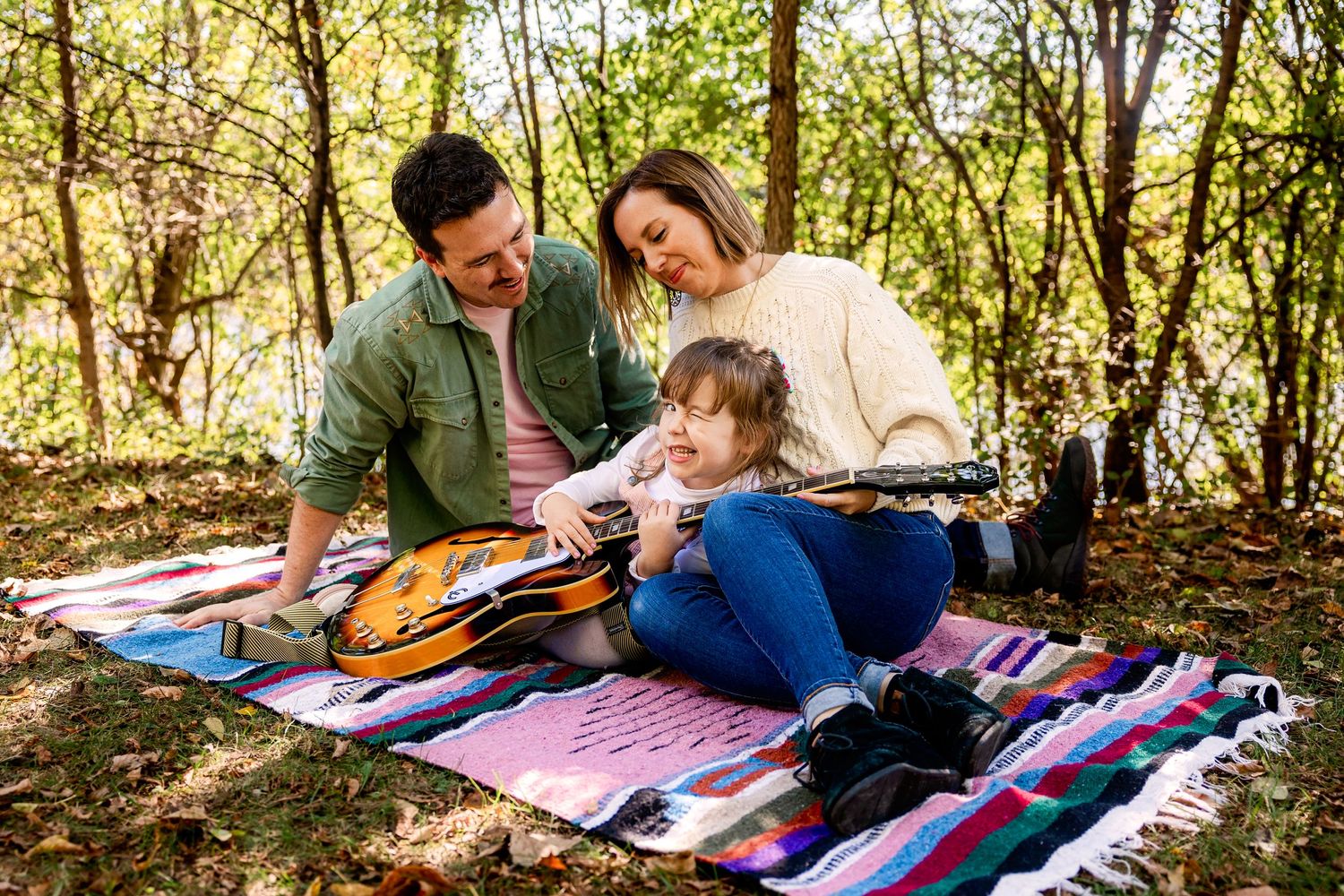 Family enjoying outdoor music time on a colorful blanket in a sunny wooded area with acoustic guitar.