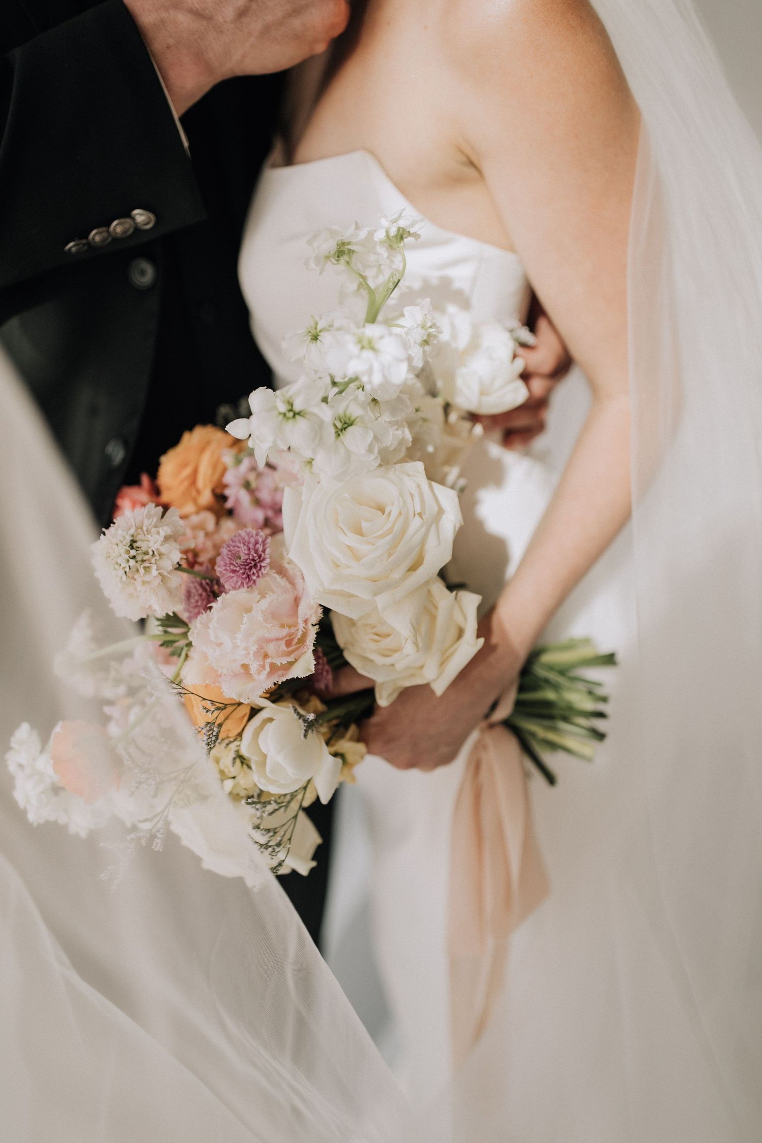Close-up of delicate white and blush wedding bouquet with roses against bride's dress and groom's dark suit jacket.