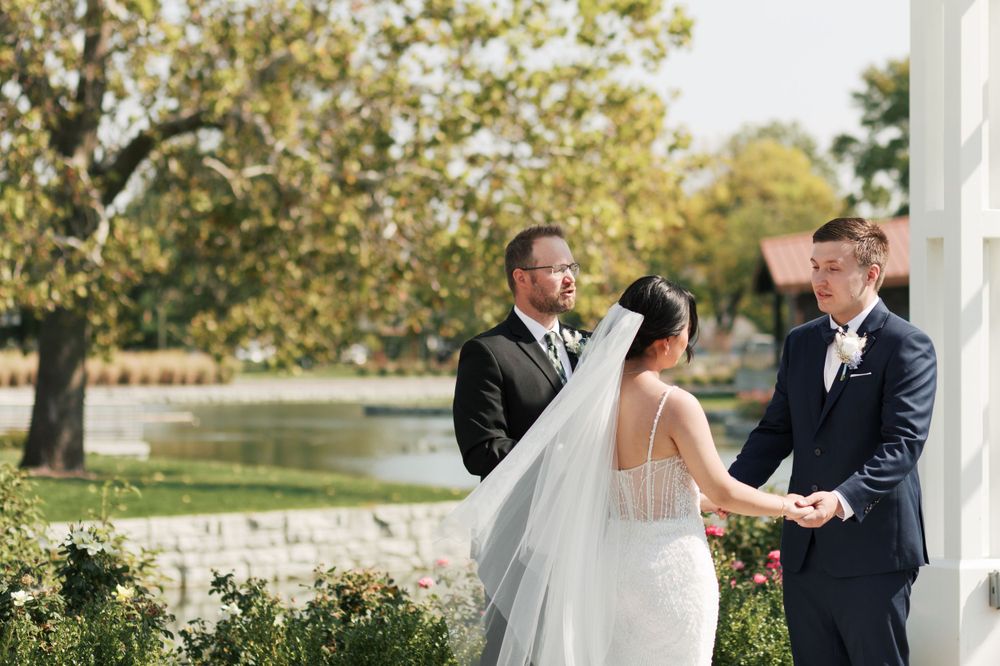 An outdoor wedding ceremony under trees with officiant and couple at decorative white altar.