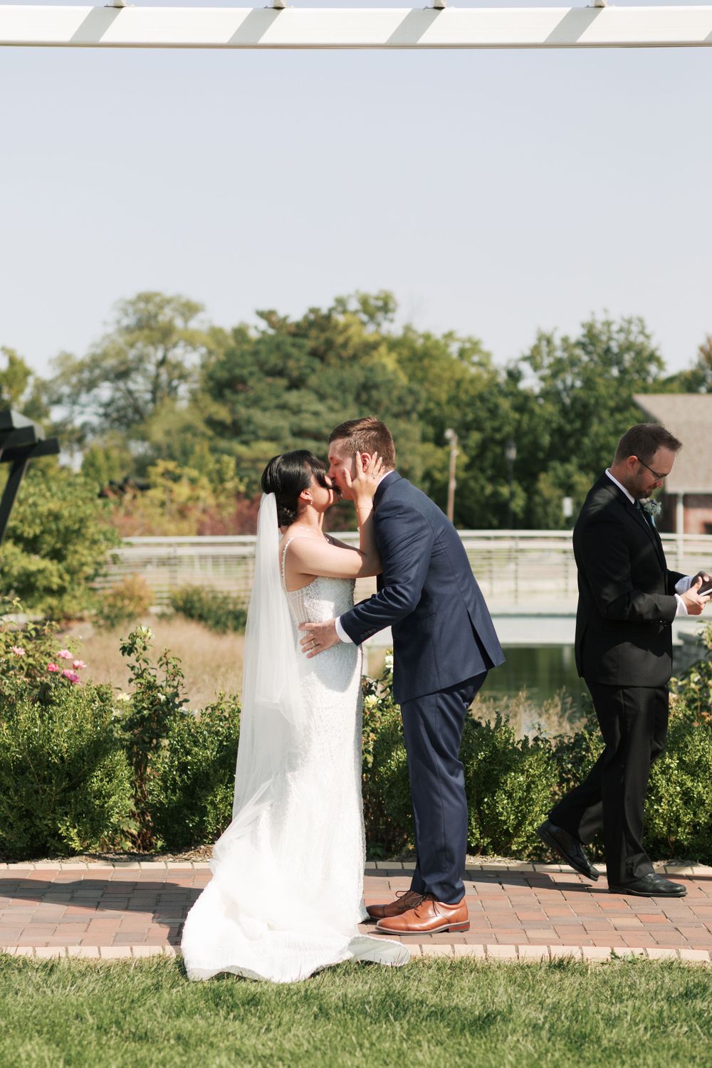 A romantic wedding kiss on a garden pathway with lush greenery and flowers in the background.