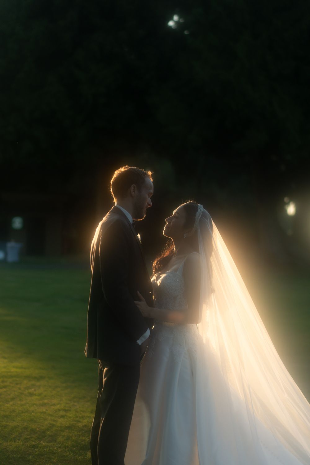 Backlit wedding couple standing on lawn with dramatic golden rim lighting.