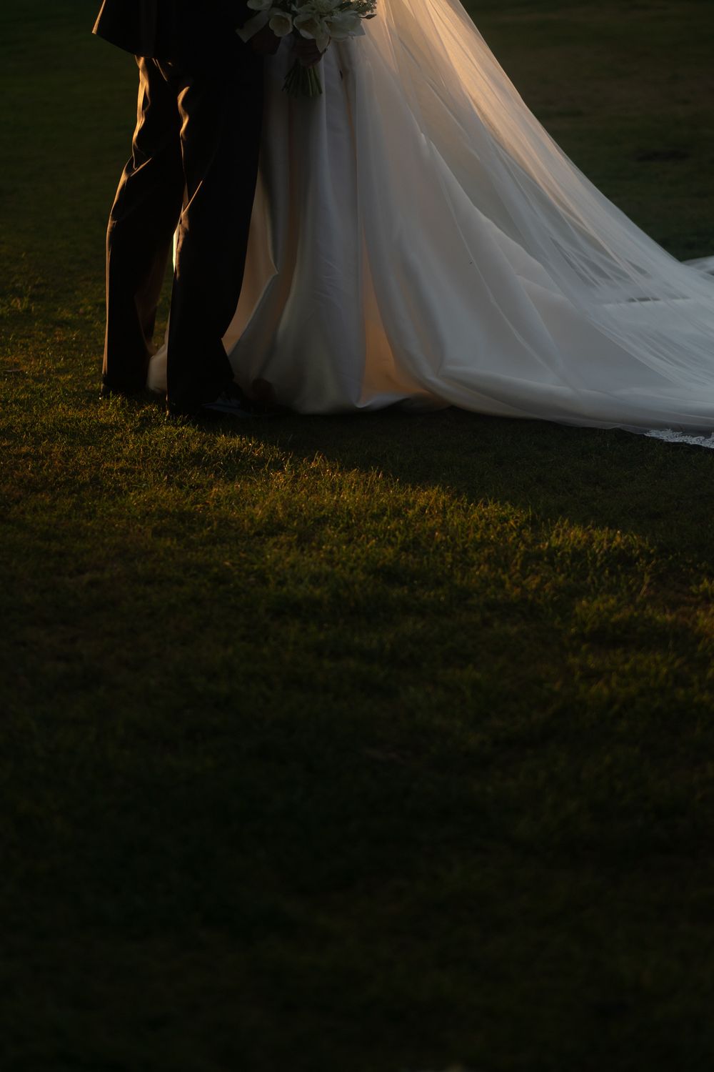 Flowing white wedding dress train and dark suit silhouetted against grass in golden light.
