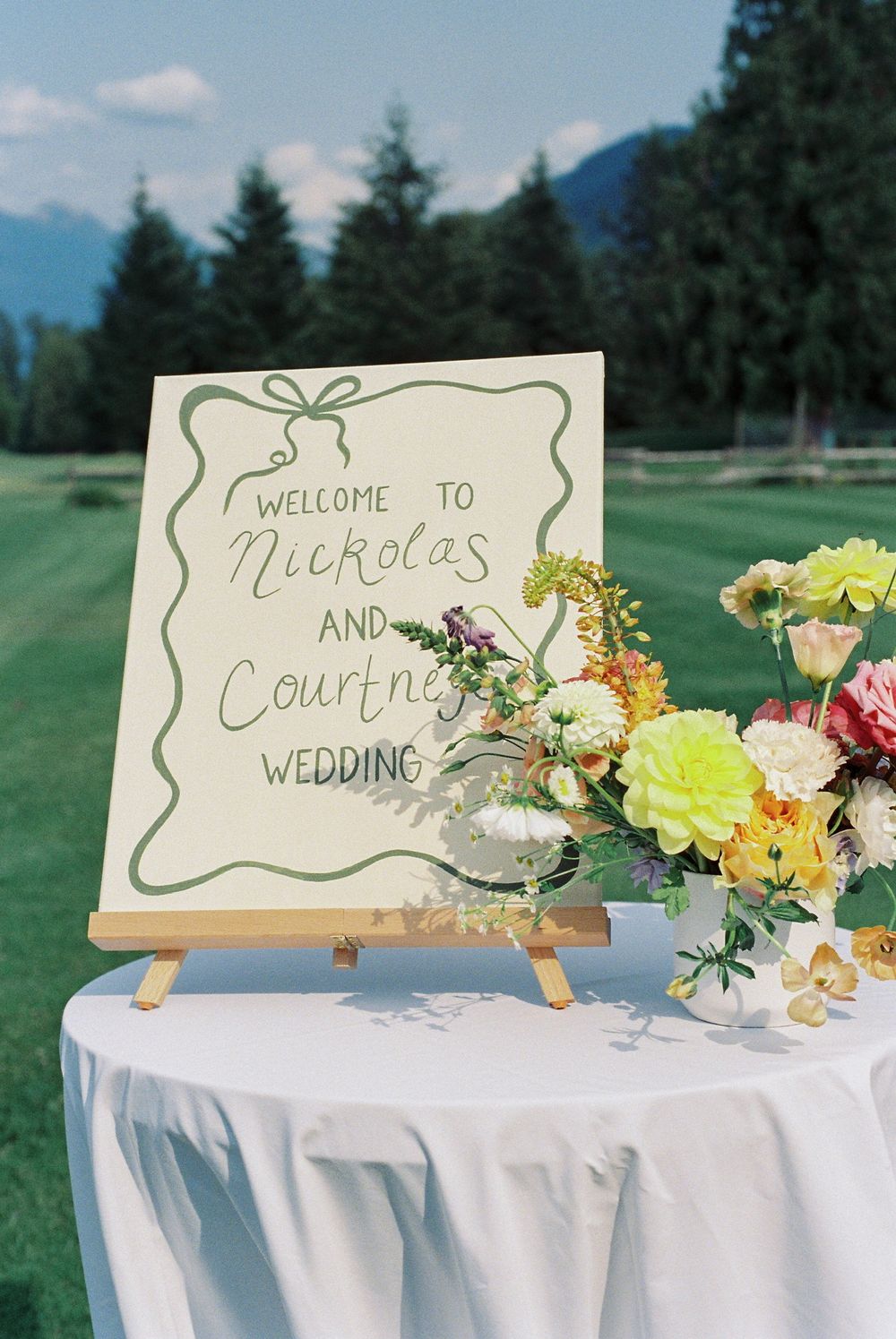 A wedding welcome sign with floral decorations displayed on an easel against a mountain backdrop.