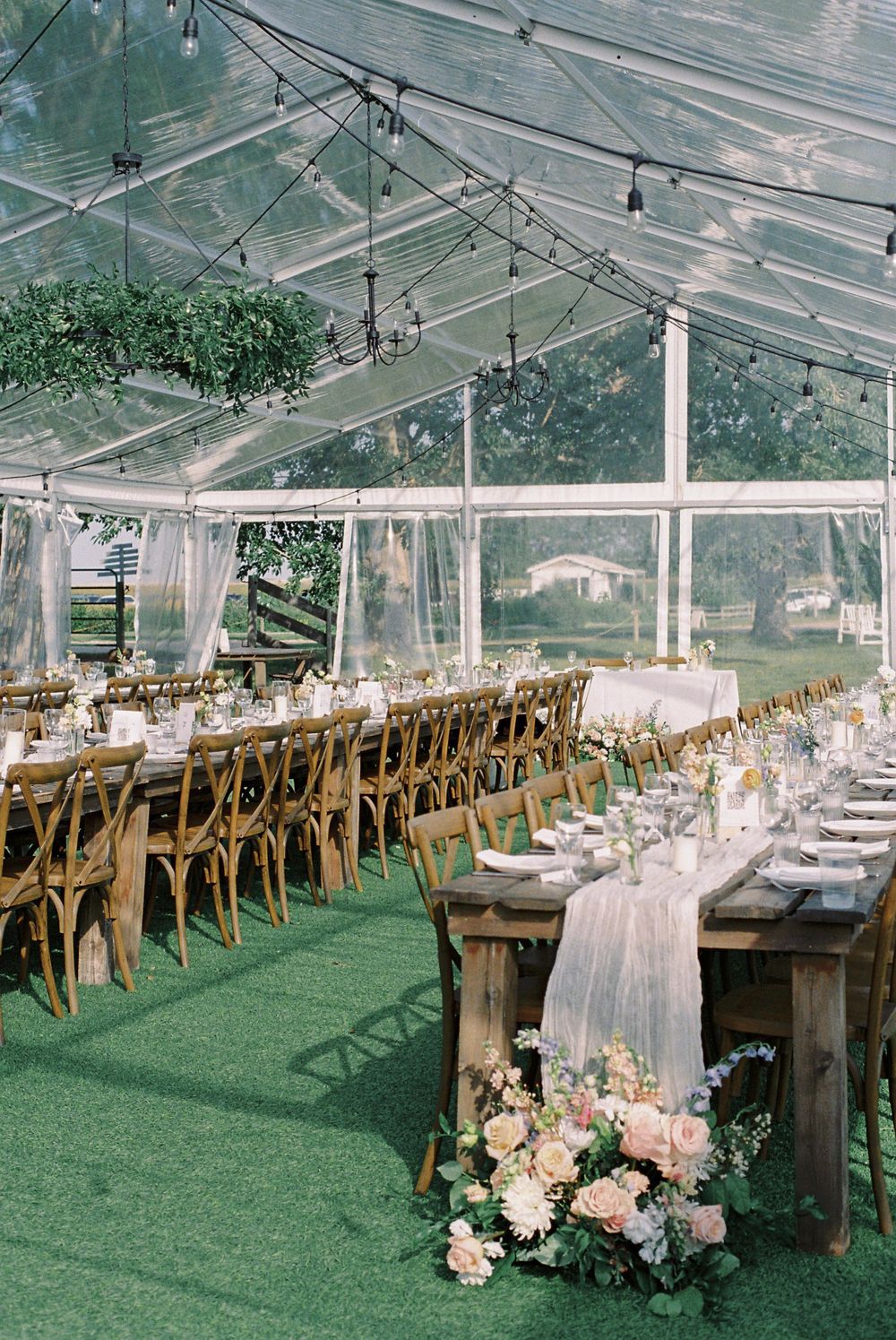 Clear tent wedding reception setup with rustic wooden tables and cross-back chairs.