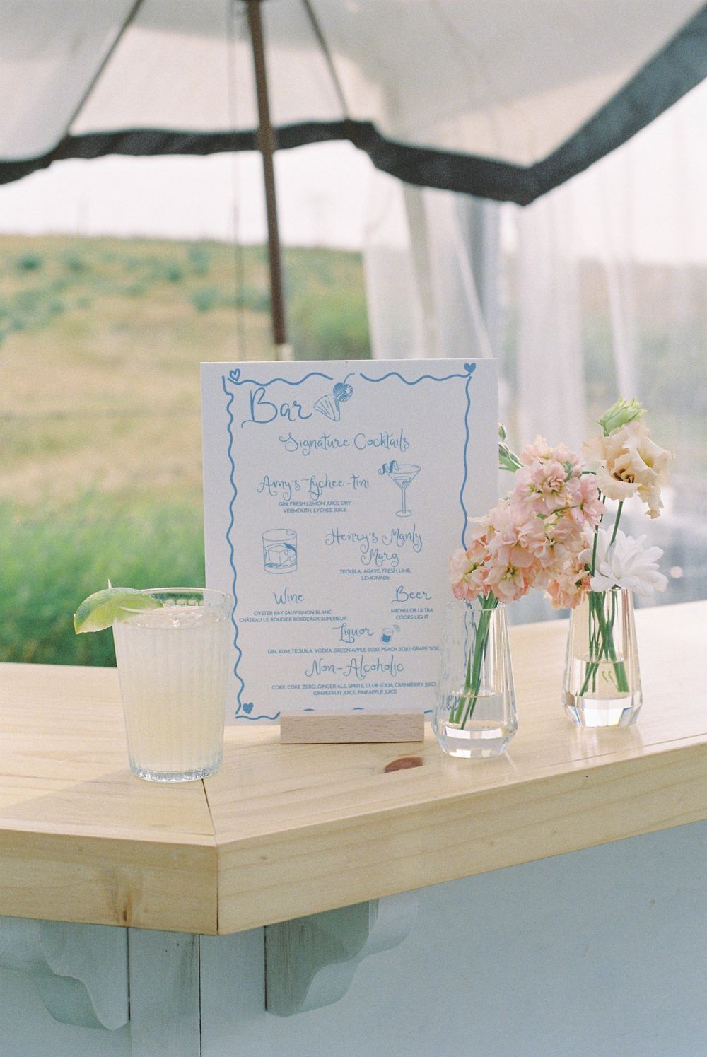 Bar menu and floral arrangements displayed on wooden table at outdoor event.