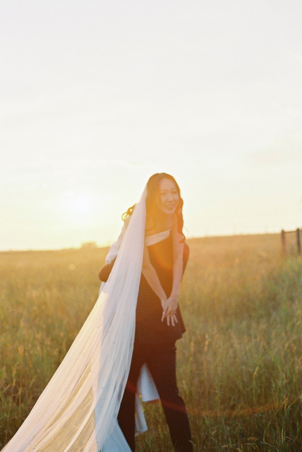 A flowing white wedding dress and veil catch the sunset light in a rustic field setting.