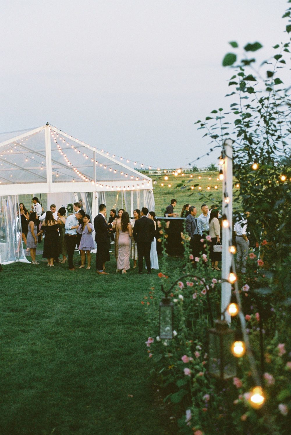 Wedding guests gather under a clear tent decorated with string lights at an outdoor evening reception.