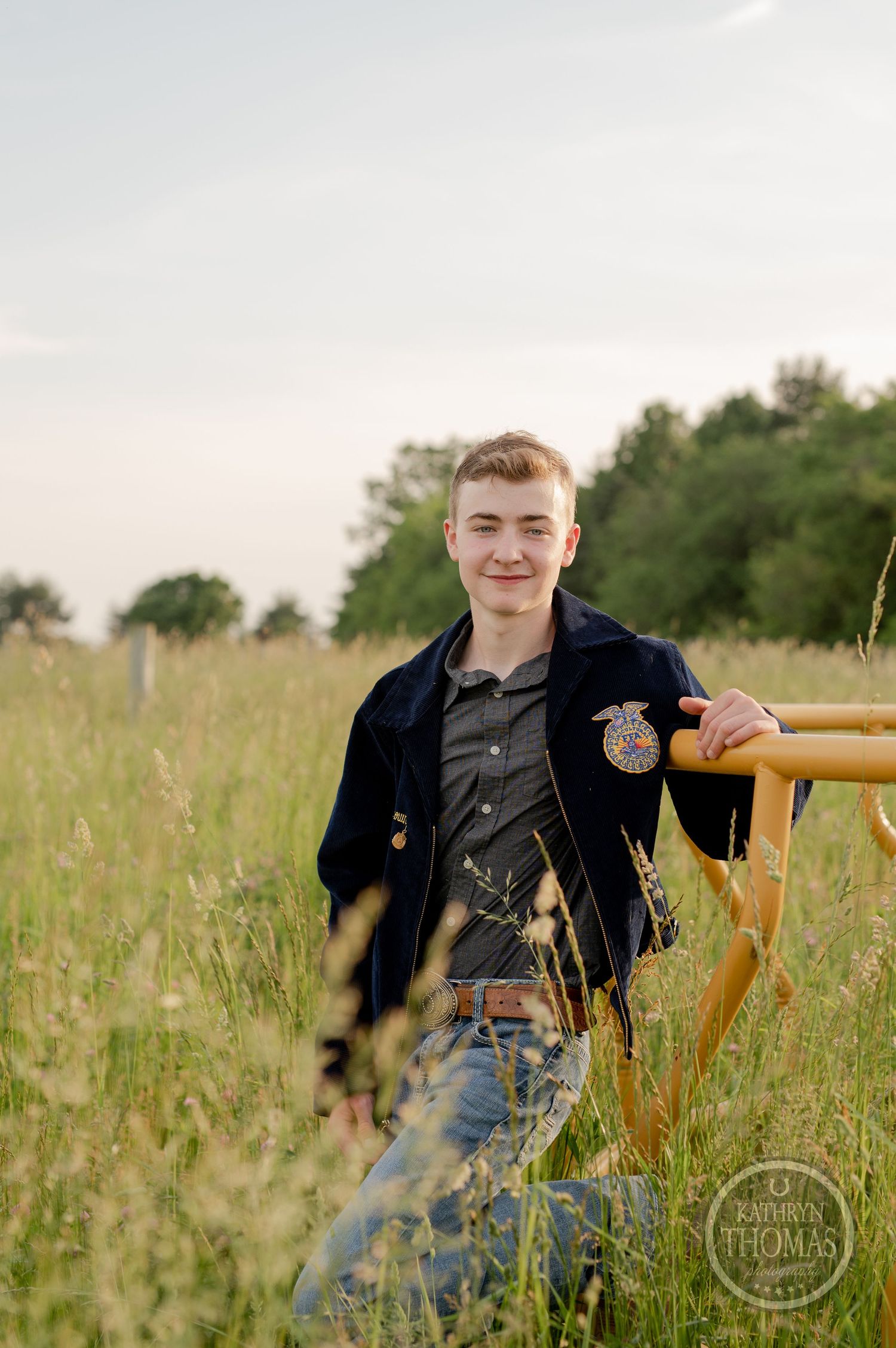 Student wearing FFA jacket poses casually against yellow railing in tall grass field.