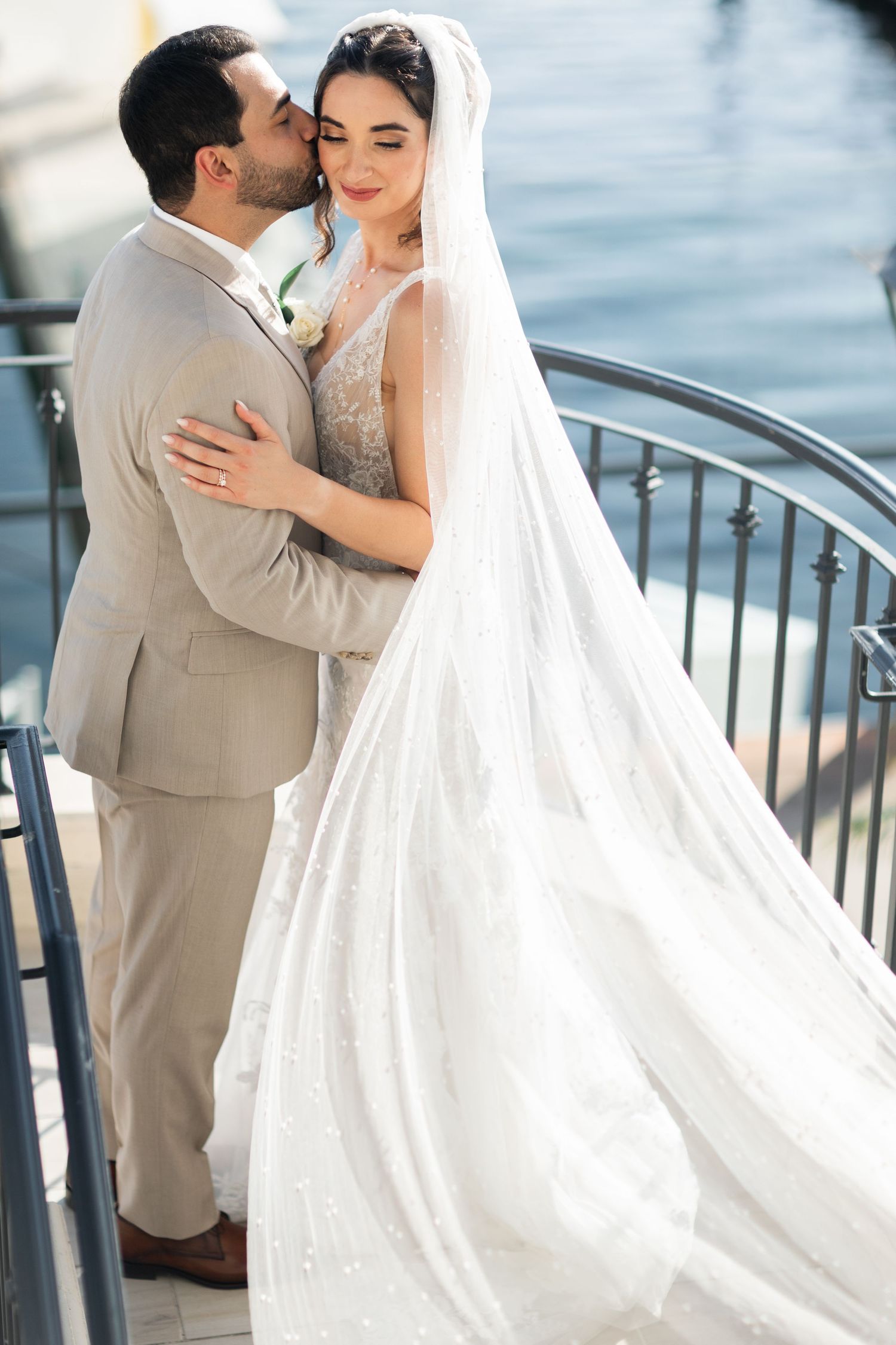 A romantic wedding couple embraces on a waterfront terrace in Newport Beach, with the bride wearing a flowing white veil and lace gown.