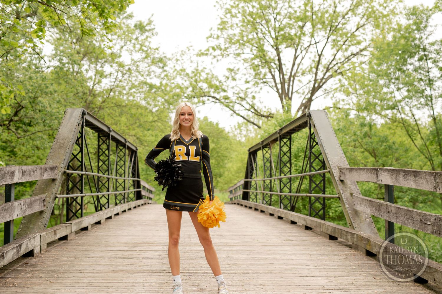 Athletic uniform-clad figure stands with pom poms on wooden footbridge among spring foliage.