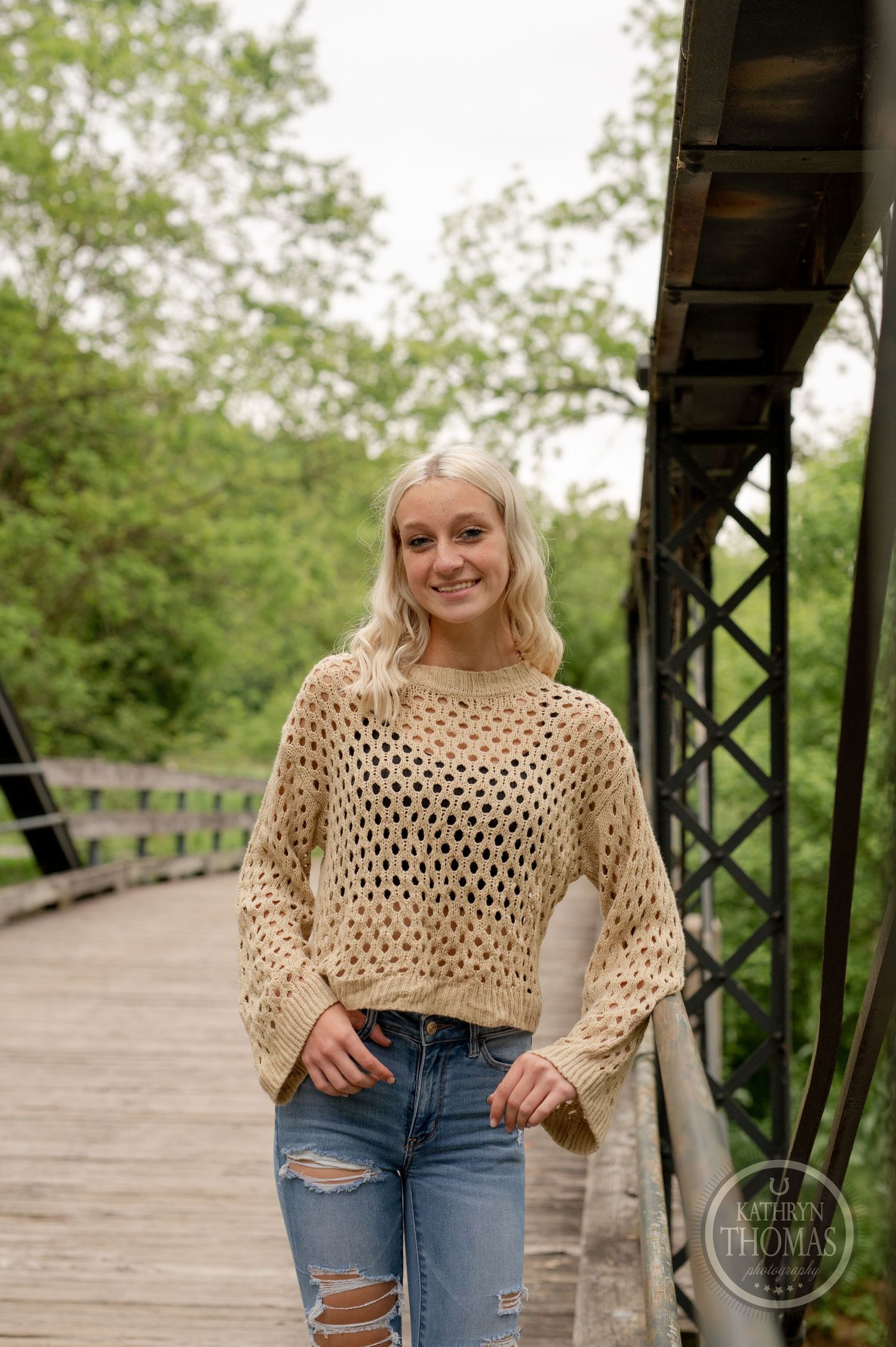 A casual spring photoshoot features a cream crochet sweater and distressed jeans on a wooden bridge.