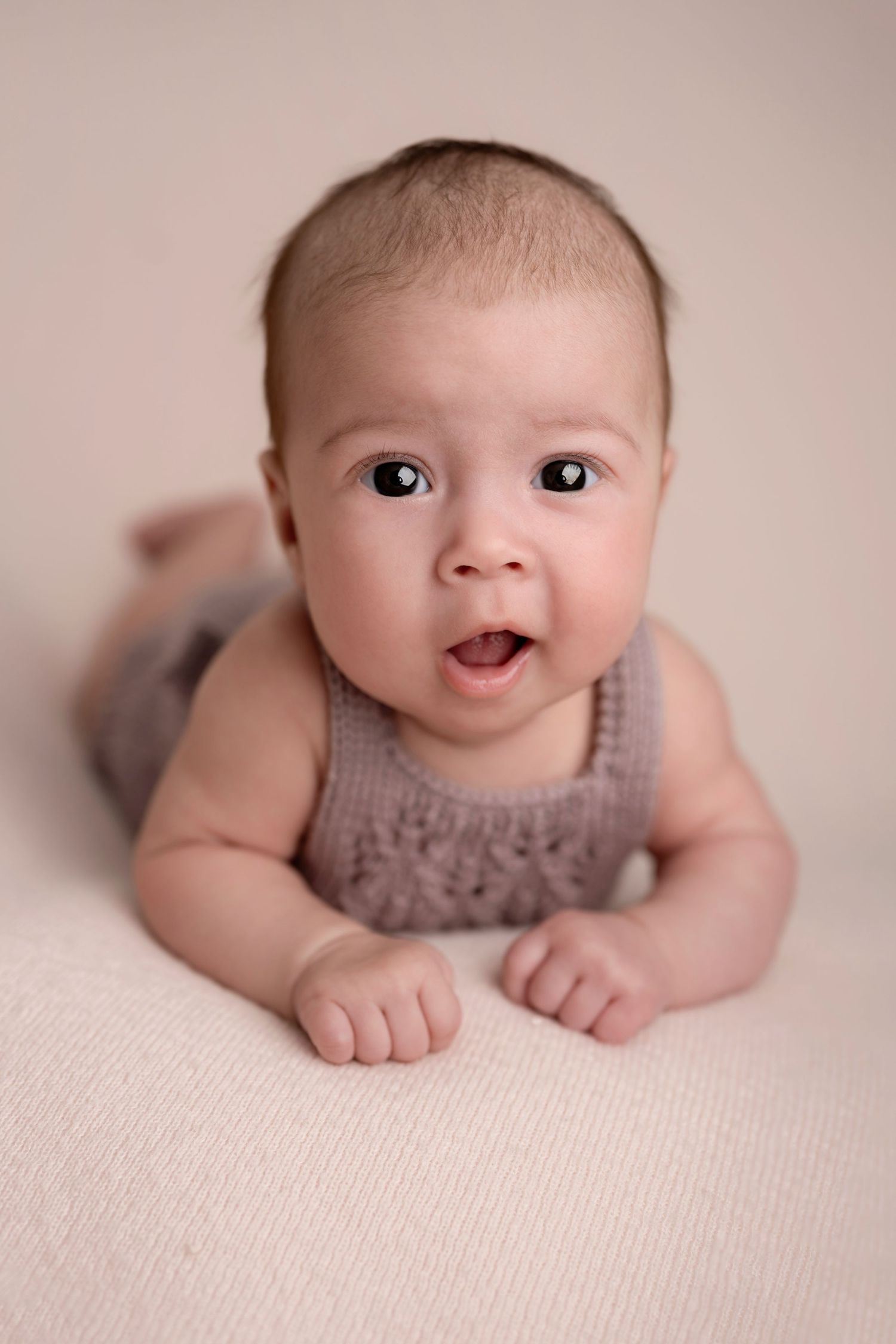 Adorable baby in a knit gray romper doing tummy time, looking up with wide eyes and open mouth on a cream blanket.