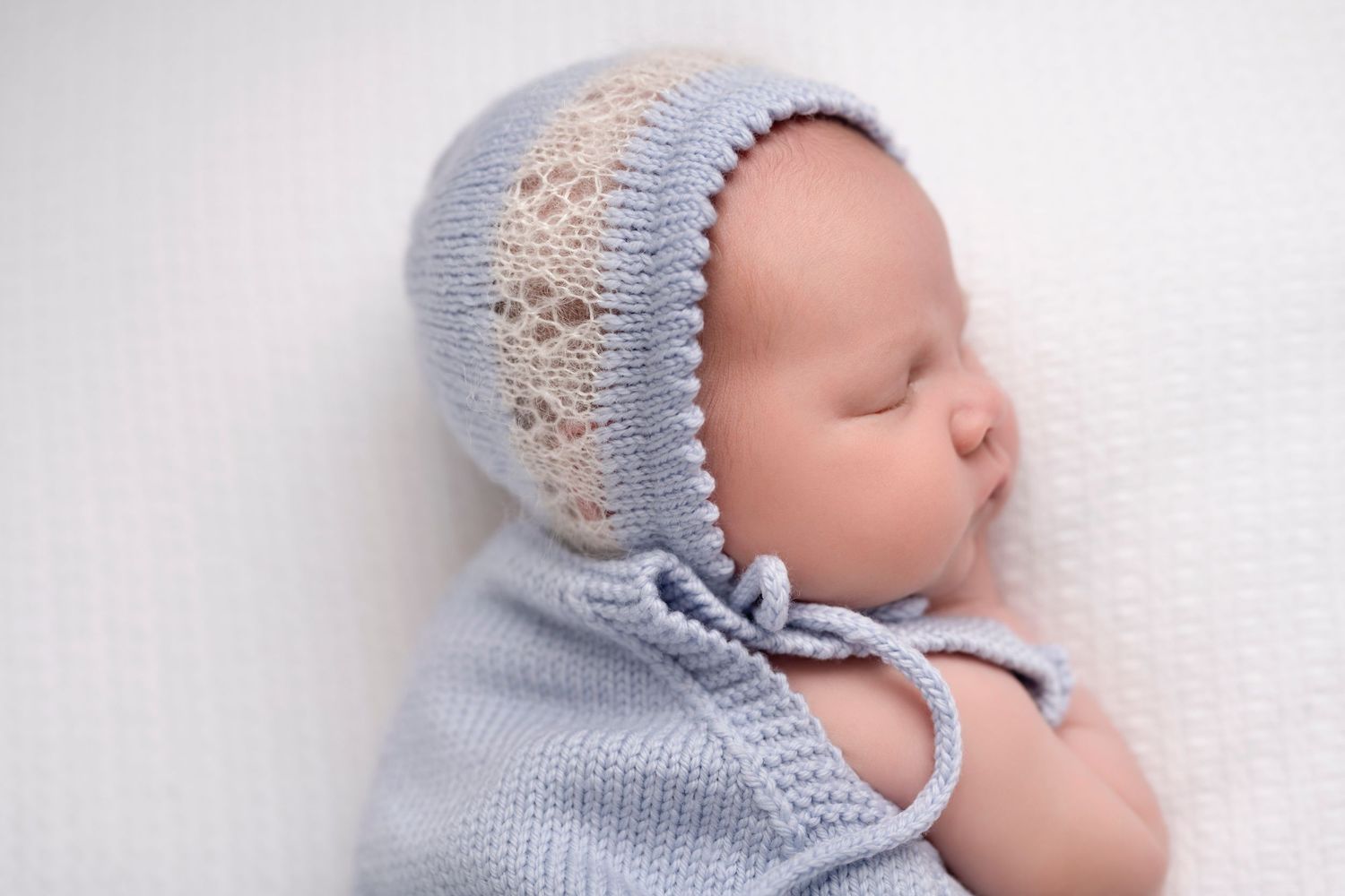 Sleeping newborn baby wearing a delicate blue knit bonnet with lace detail, wrapped in a soft blue blanket.
