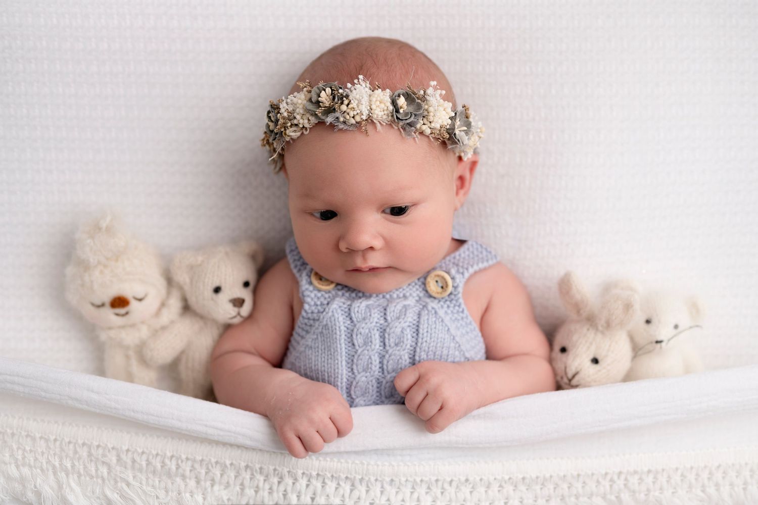 Newborn baby wearing a floral headband sits surrounded by small white teddy bears on a white knit blanket.