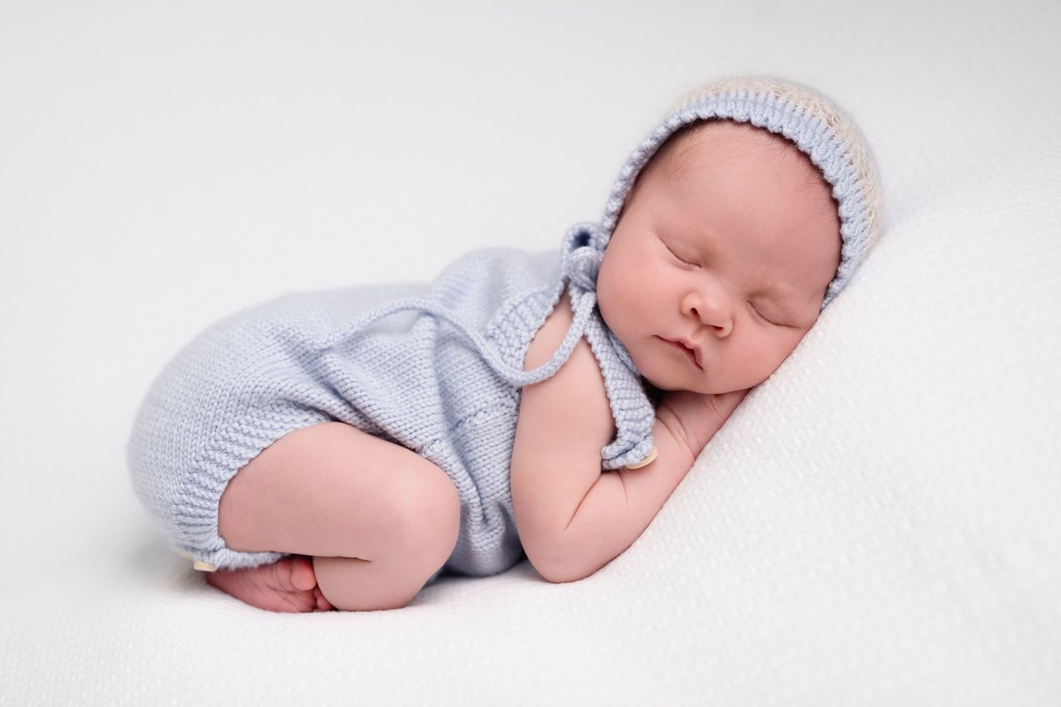 Peaceful newborn baby sleeping in blue knit romper and bonnet on a soft white blanket.
