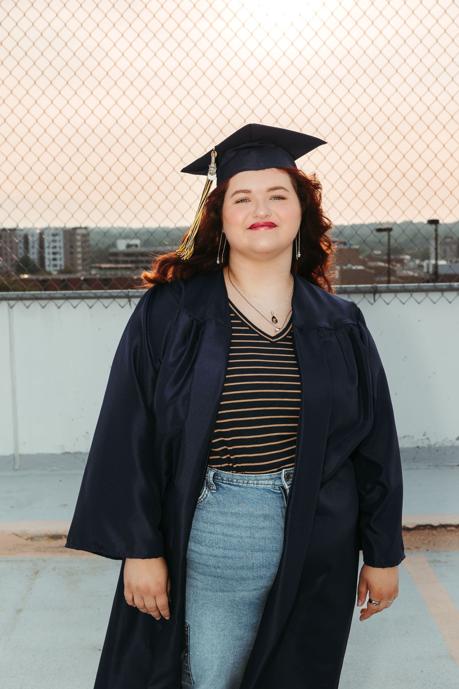 Graduate student wearing a cap and gown poses outdoors on a rooftop against an urban backdrop at sunset.