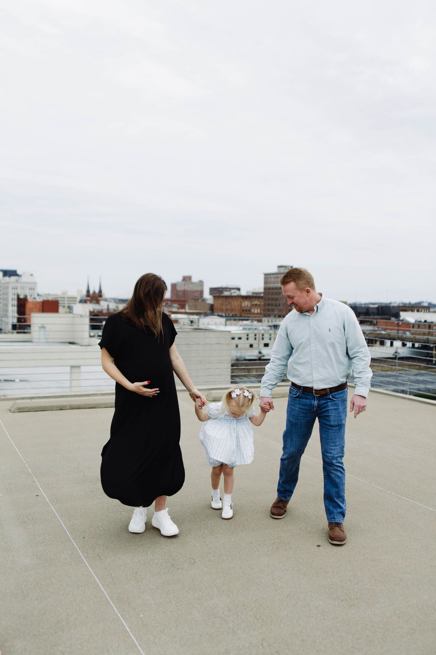 Family holding hands with small child in white dress while walking on urban rooftop.