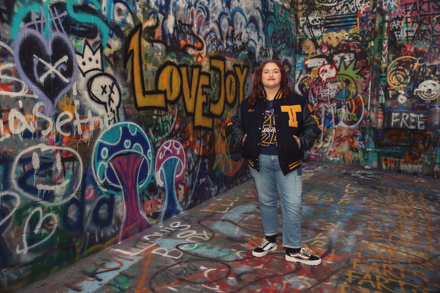 A high school senior stands against a vibrant graffiti wall wearing a denim jacket and jeans in an urban setting in Downtown Ann Arbor.