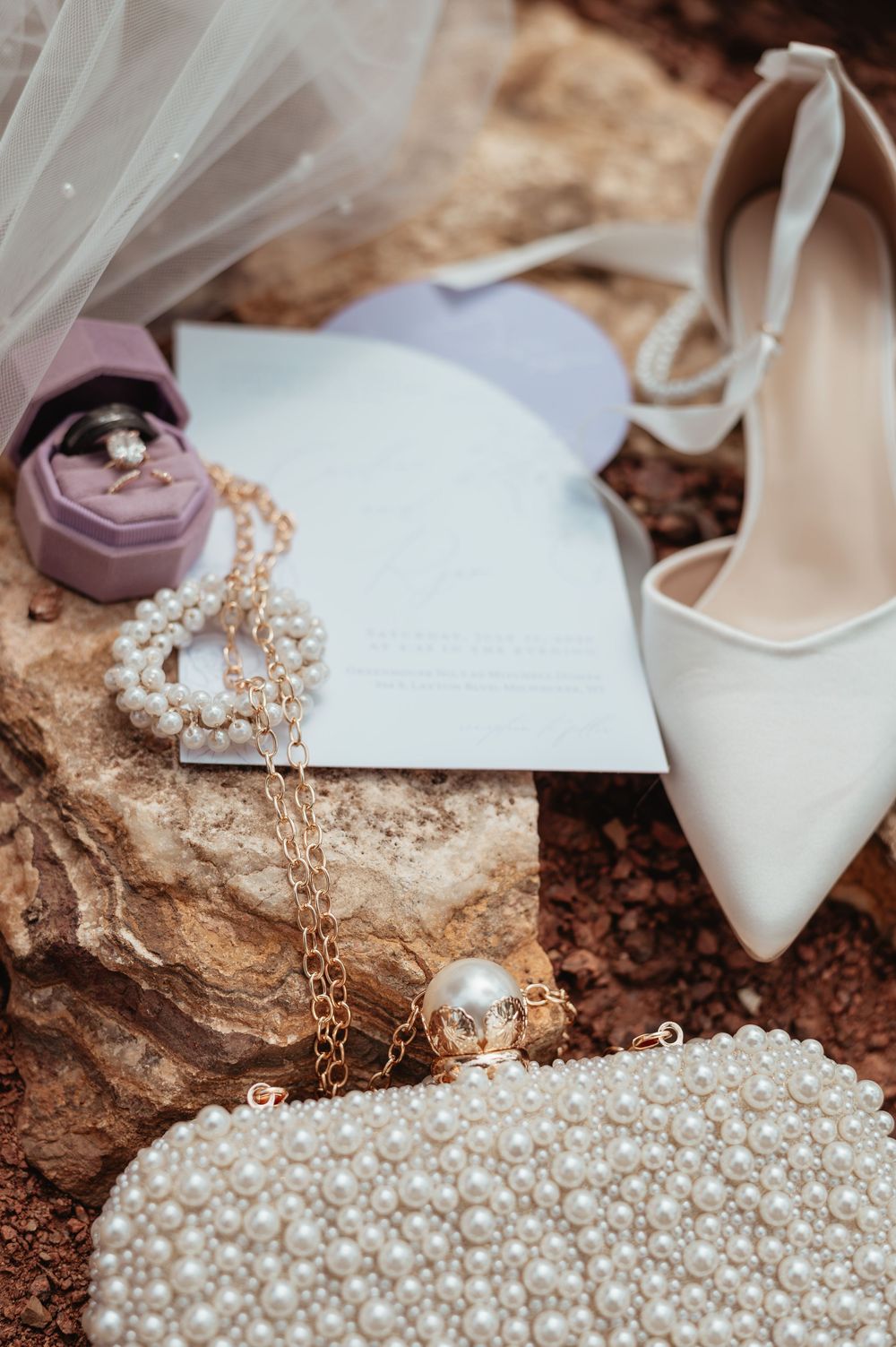 Elegant bridal flat lay with pearl clutch, white heels, jewelry, ring box, and wedding invitation on rustic stone.