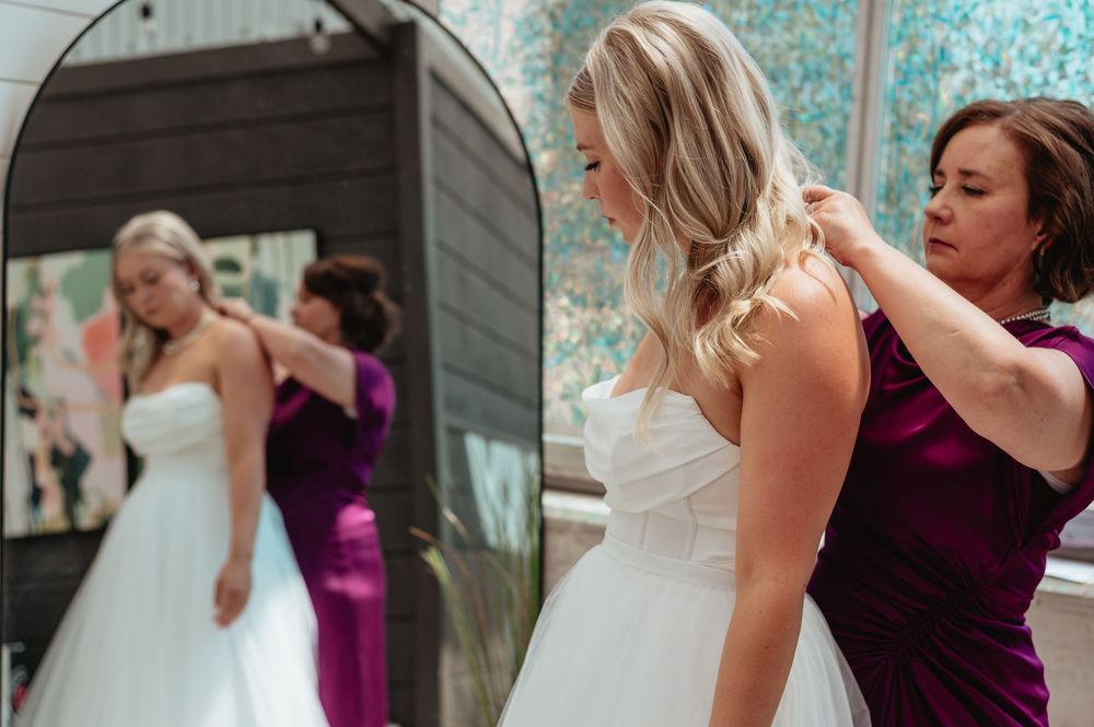 Bridesmaid in purple dress helping bride with white wedding gown while reflections show in mirror.